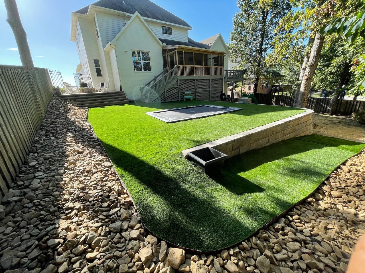 Backyard with artificial turf, retaining wall, house, rocks, and a trampoline. Sunny day.