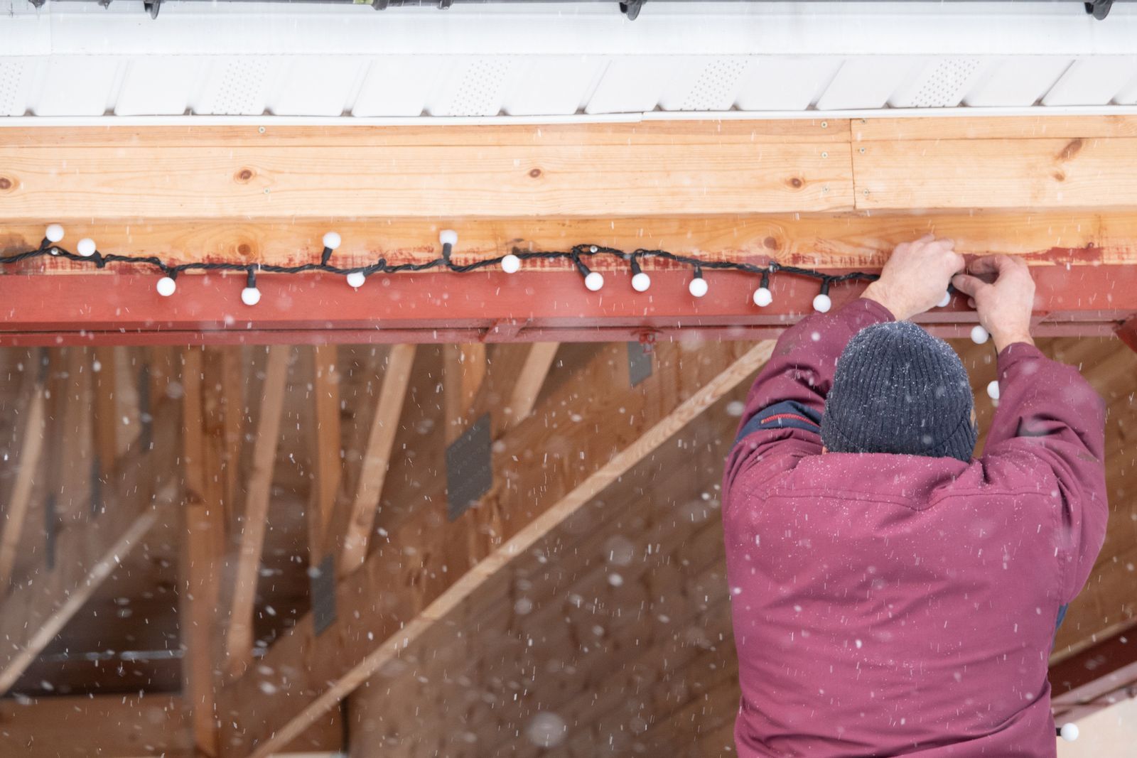Person in maroon coat hangs Christmas lights on wooden structure in the snow.