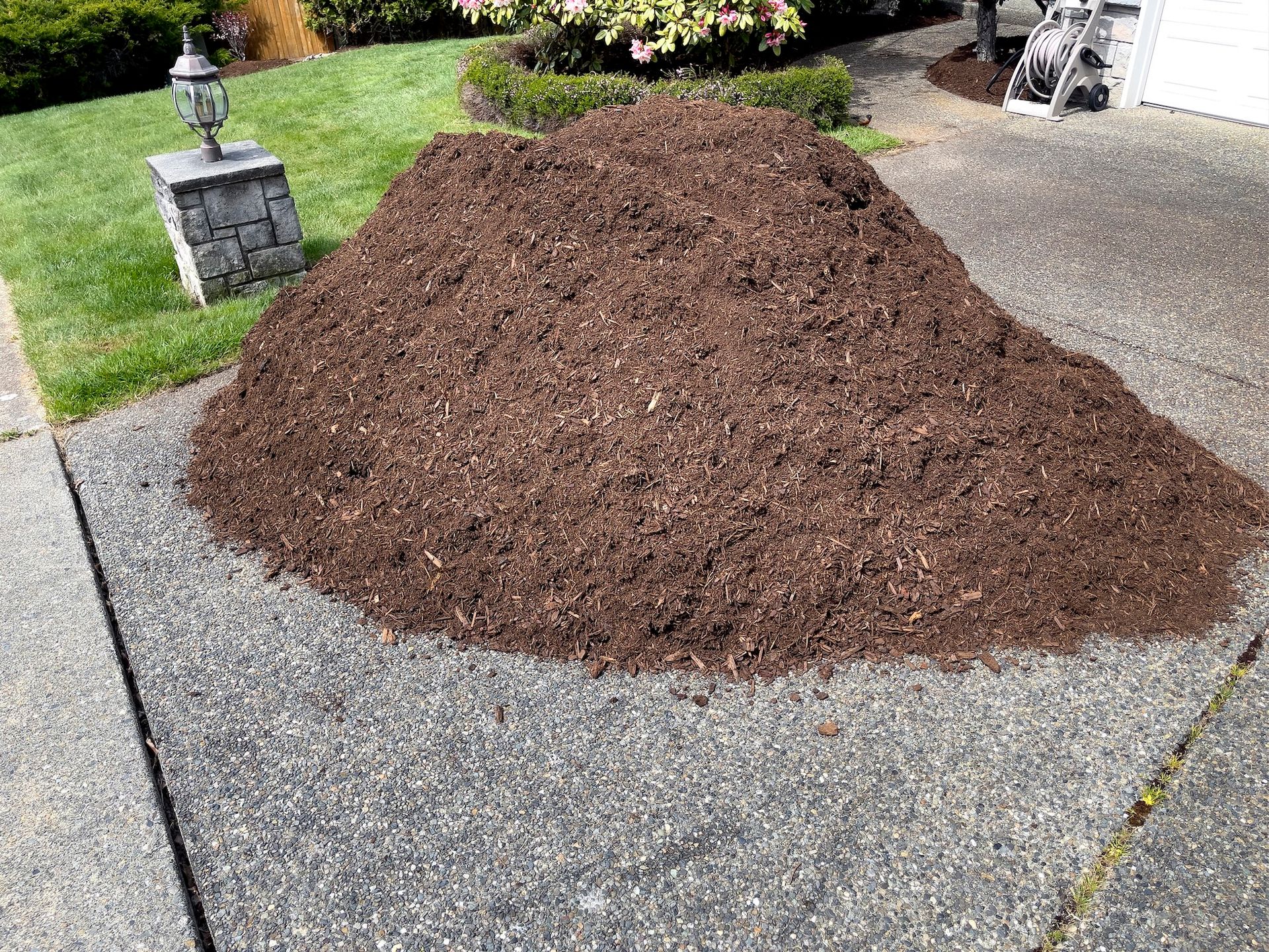 Pile of brown mulch on a gray concrete driveway, near a patch of green grass and a garden.