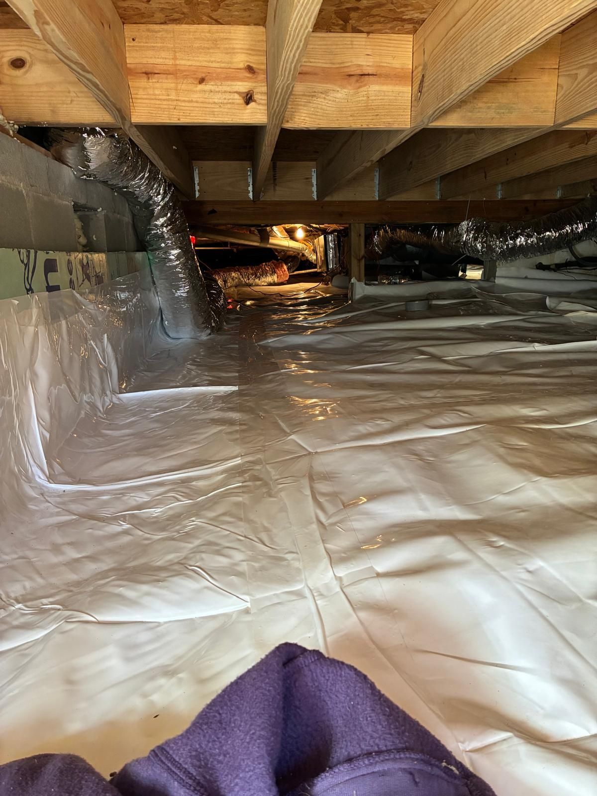 Crawlspace with exposed wooden joists and plastic vapor barrier, lit by a dim light in the distance