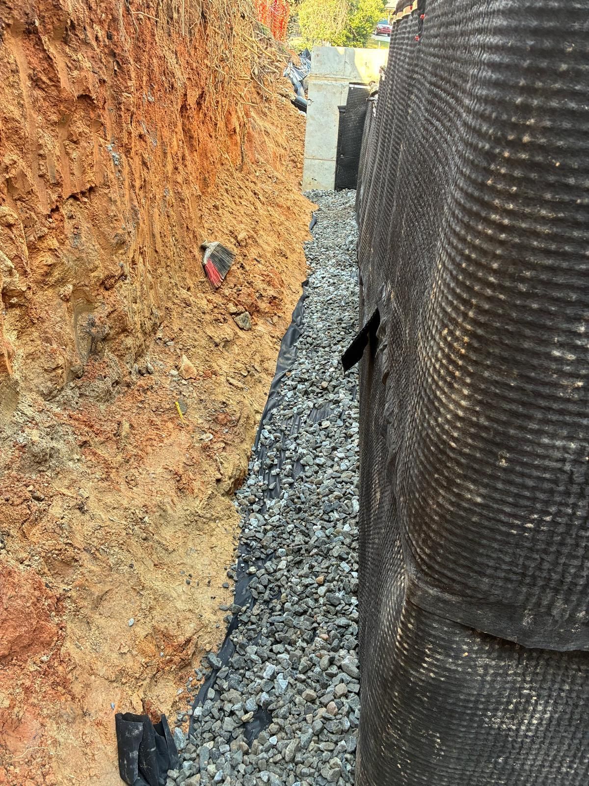 Narrow gravel trench between an excavated dirt wall and a black drainage pipe or liner