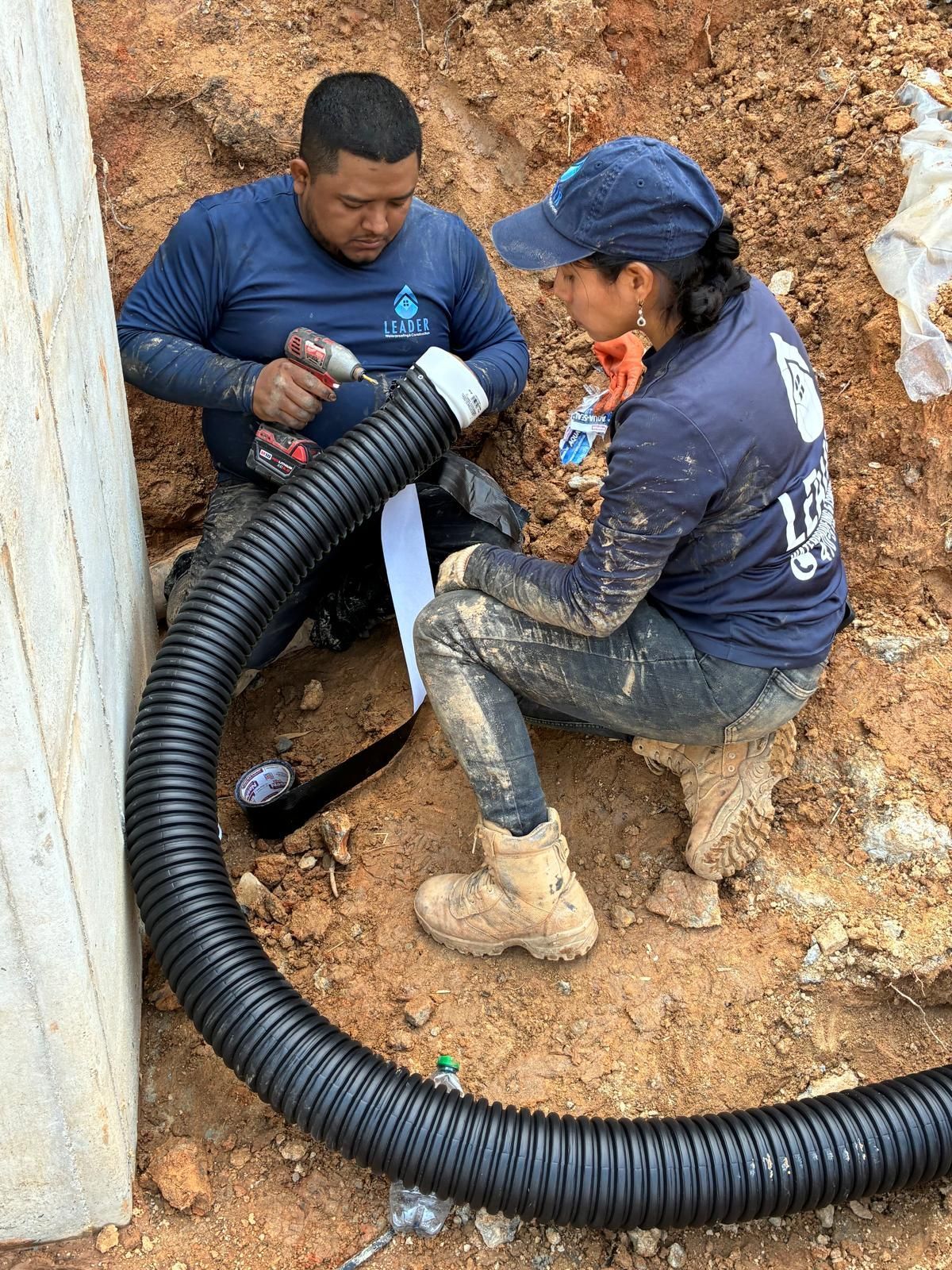 Two workers connect a large black corrugated pipe in a muddy trench.
