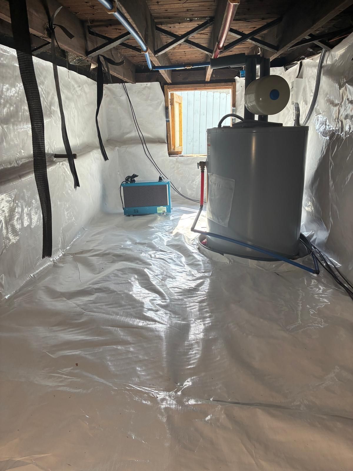 Crawlspace with plastic floor and insulation, showing a water heater, fan, and a small blue dehumidifier.