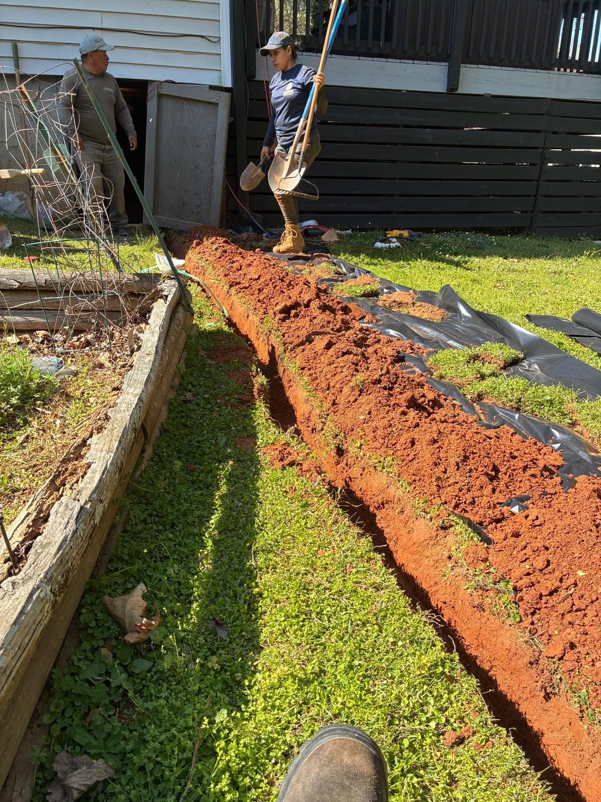 Two workers leveling a long strip of red soil beside a house and garden bed