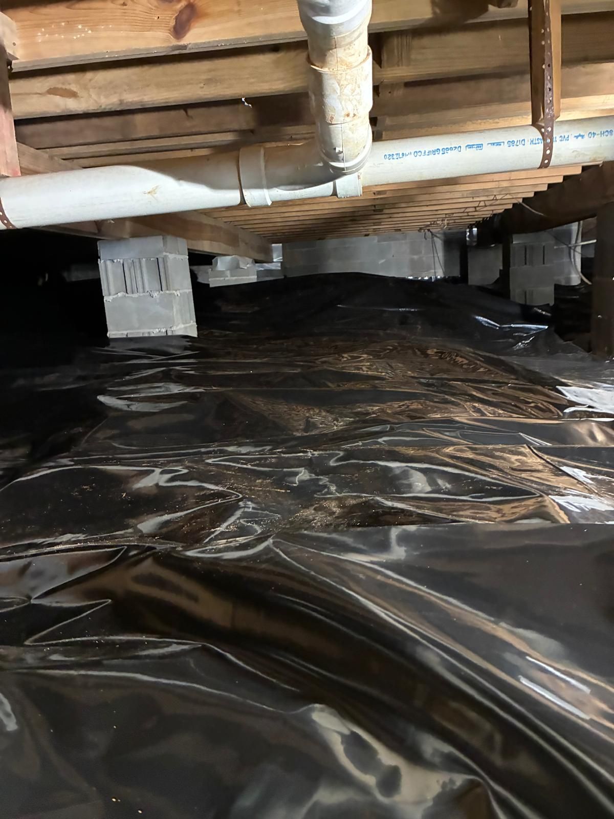 Plastic vapor barrier covering the ground beneath exposed floor joists and white pipes in a crawlspace