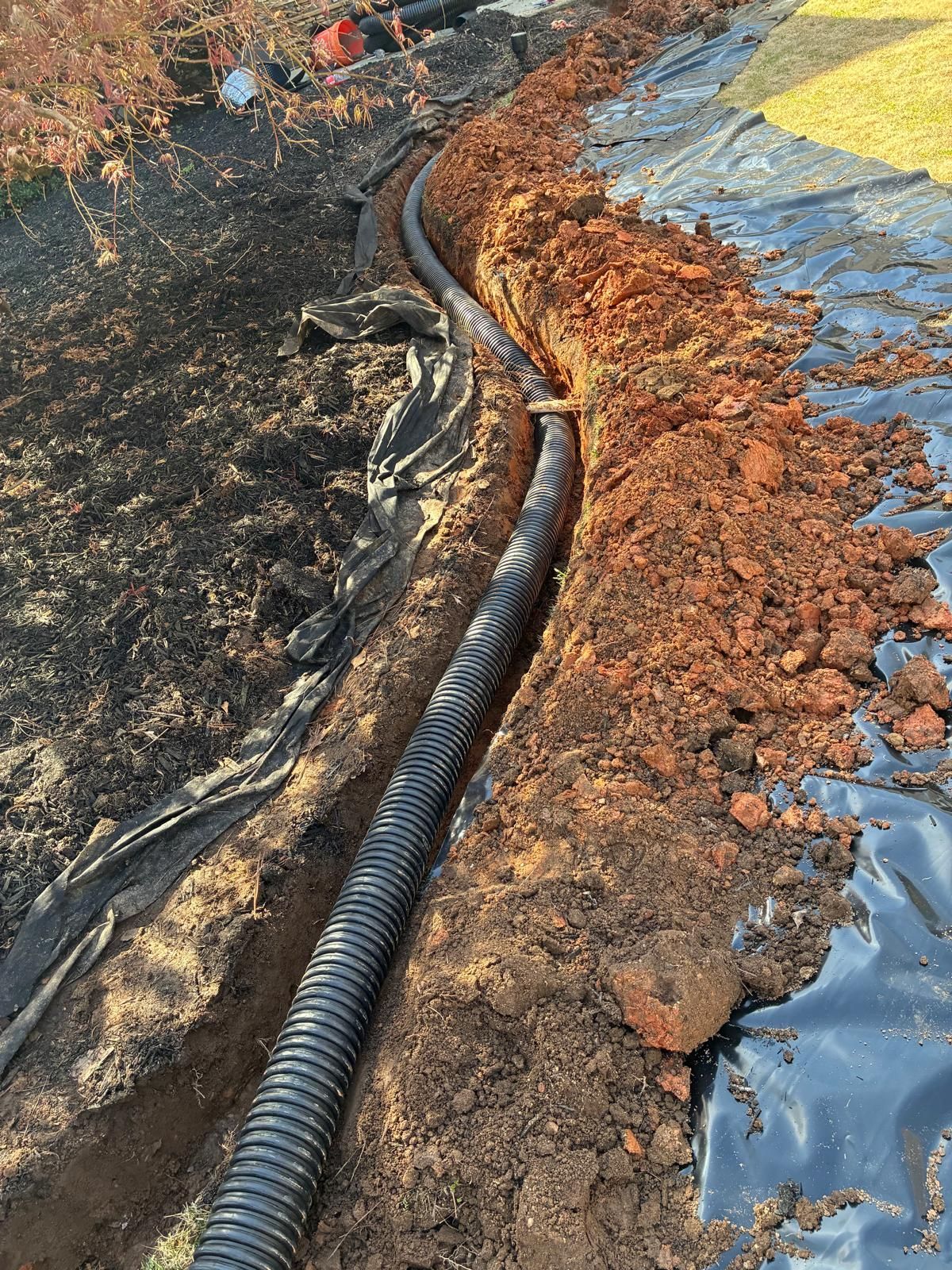 Black drainage pipe laid in a muddy trench beside a blue tarp-covered dirt embankment.