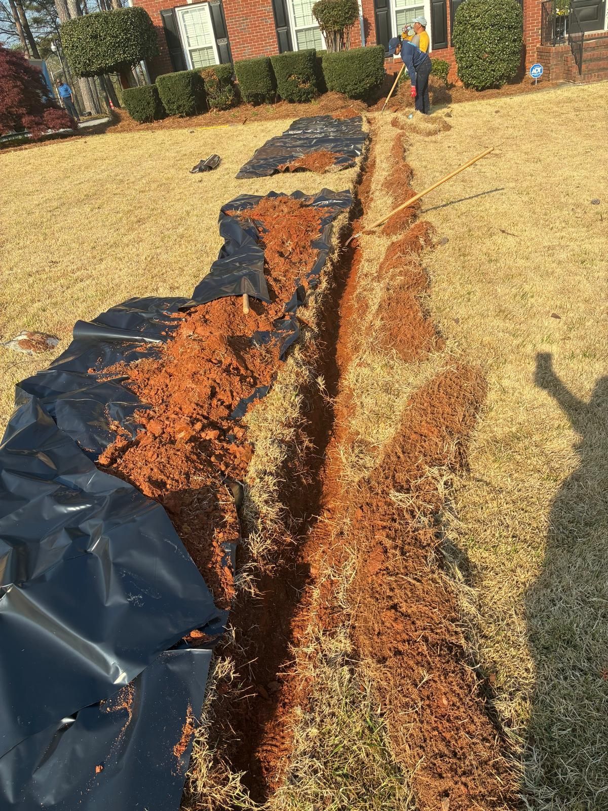 A long trench beside a house lined with black plastic and orange-brown soil, with a person at the far end.