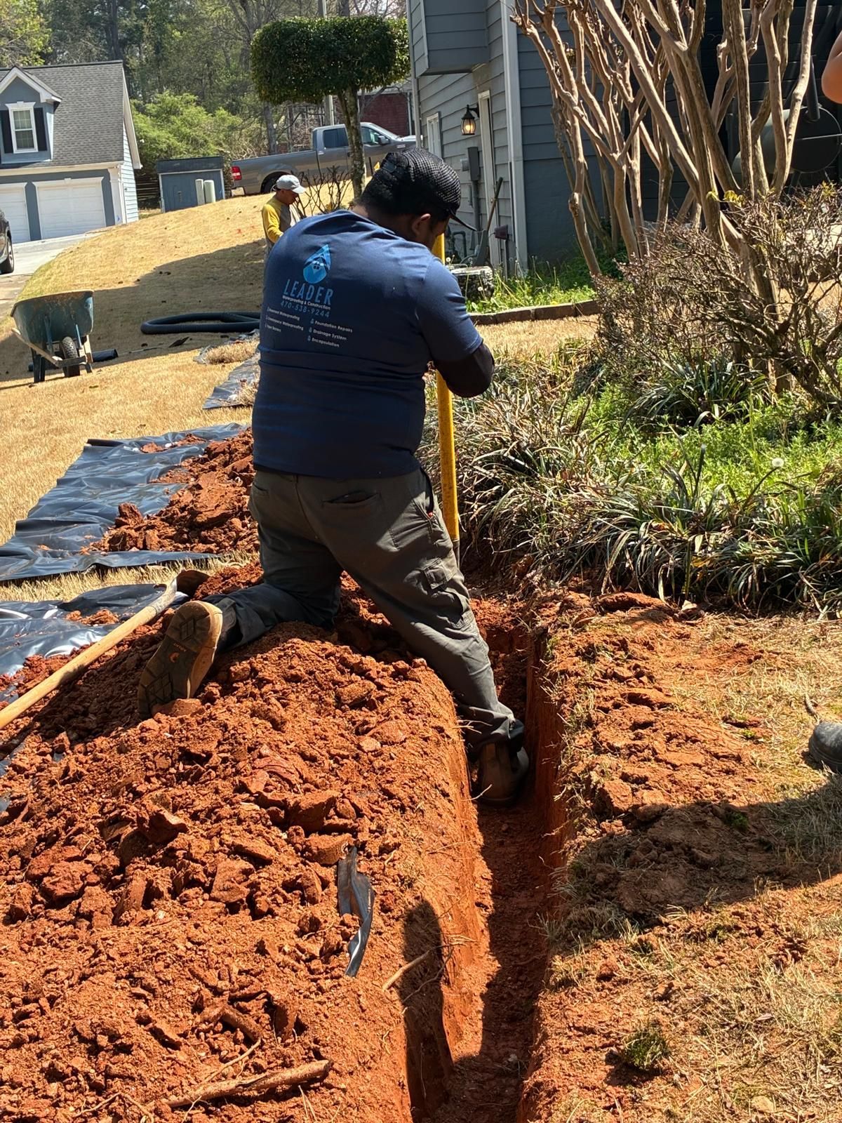 Worker digging a trench beside a house, with a yellow post hole digger and red soil nearby