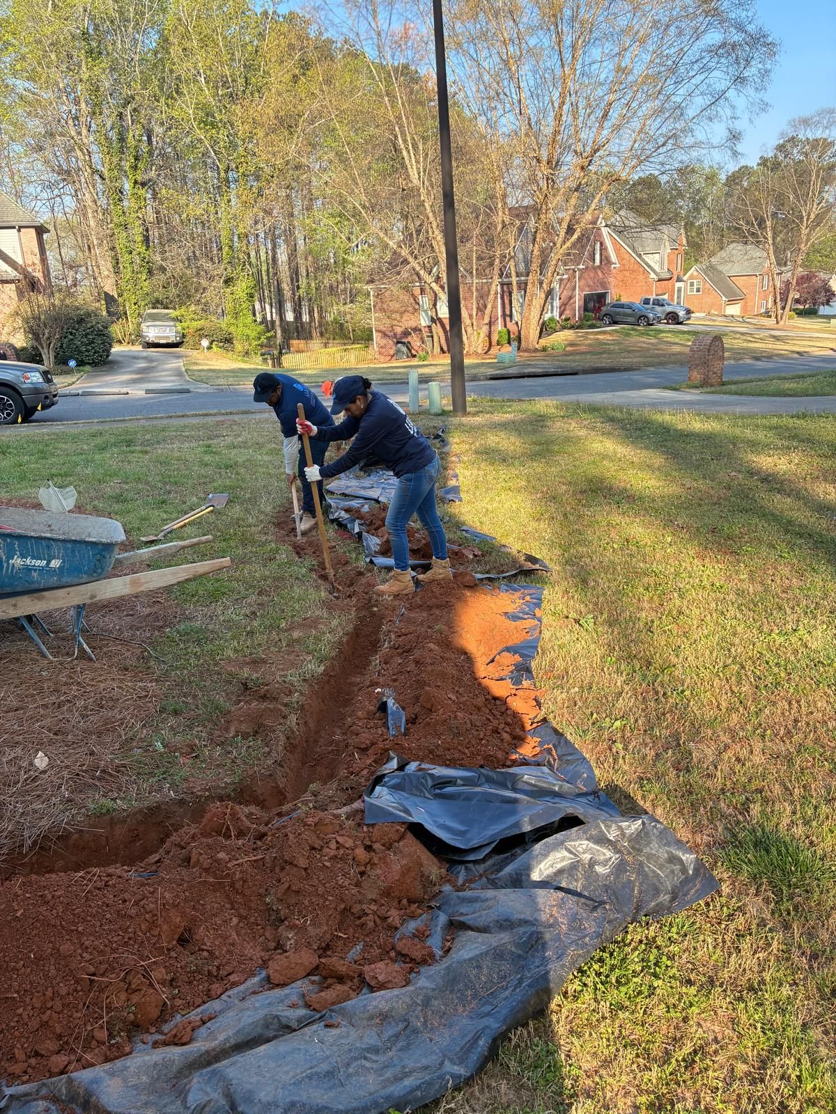 Two workers dig a trench beside a suburban sidewalk, with black landscape fabric and red soil exposed.