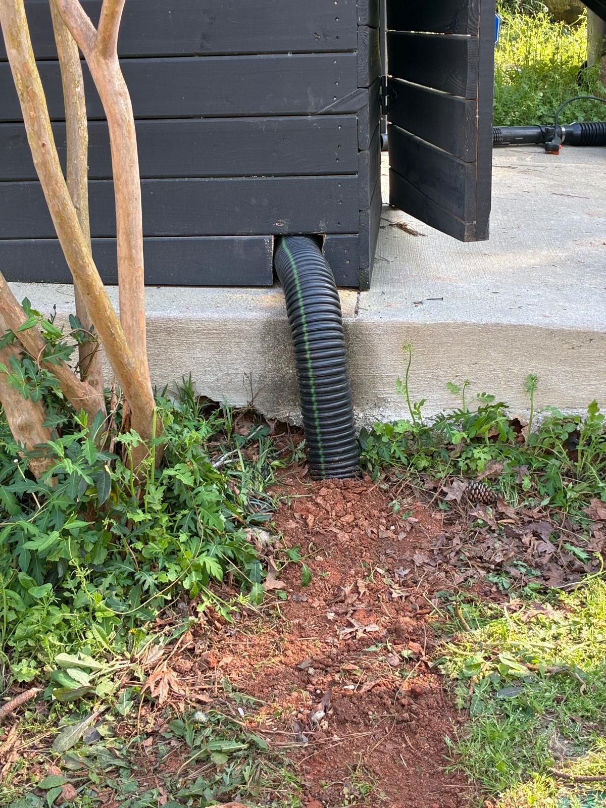 Black corrugated drainpipe under a dark shed wall beside a concrete slab and garden path