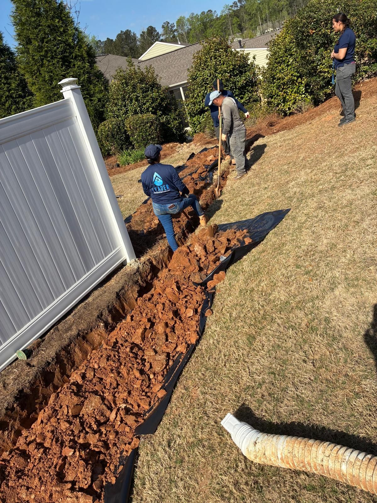 Workers digging a long trench beside a white fence on a sloped grassy yard