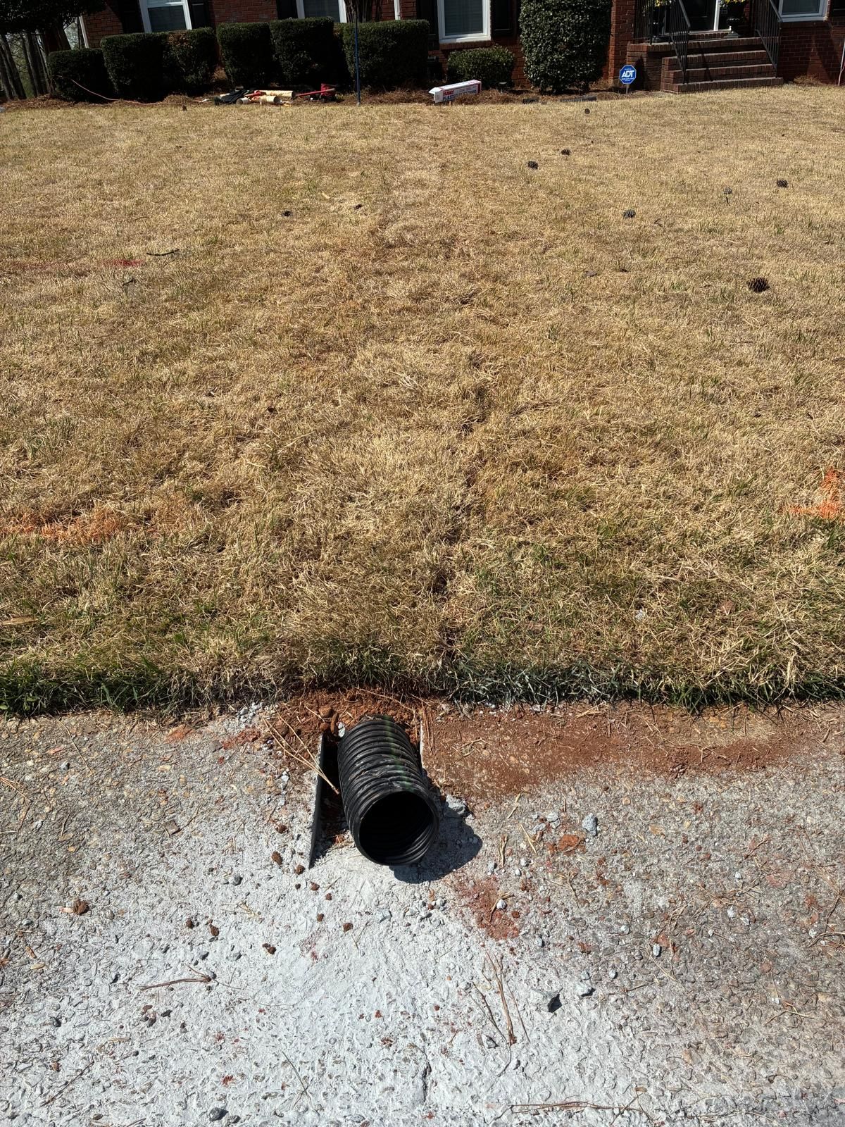 Broken black pipe opening on a concrete curb beside a dry brown lawn.