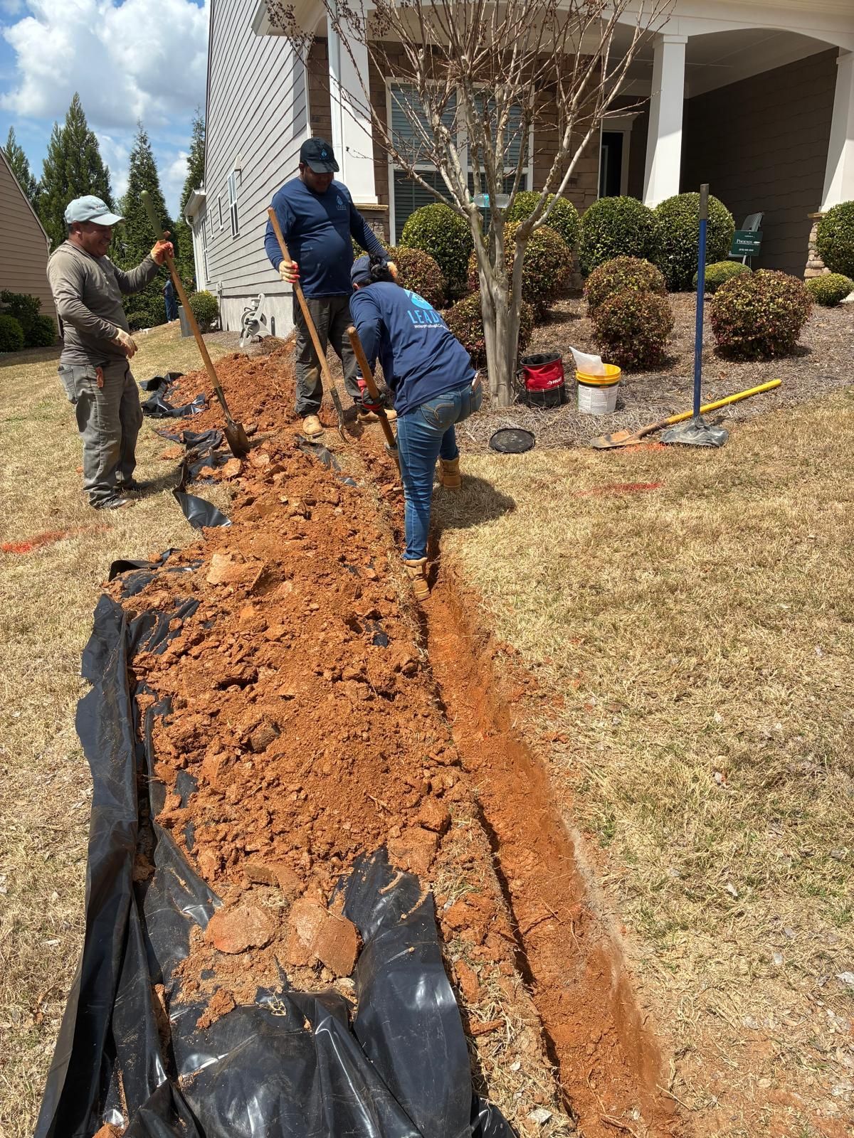 Workers digging a long trench in a front yard beside a house, with tools and black lining visible