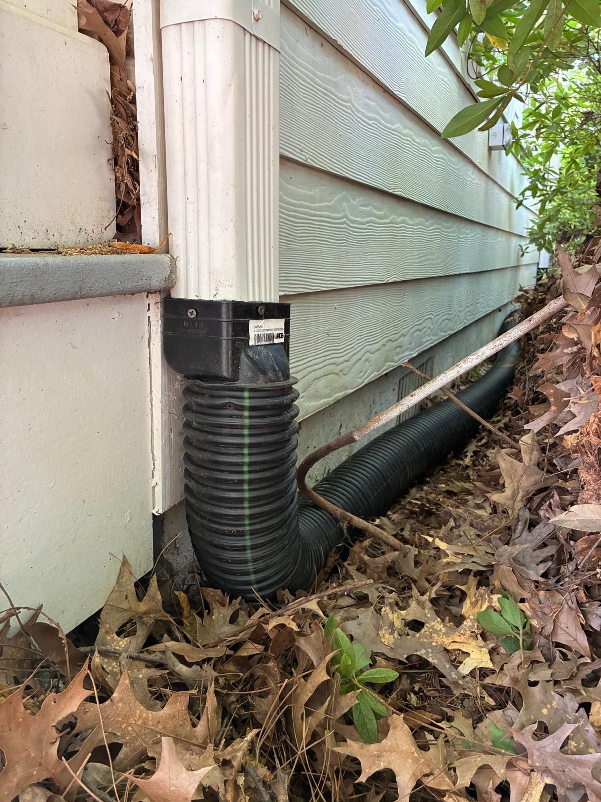 Black corrugated drainpipe beside a house foundation, surrounded by leaves and mulch