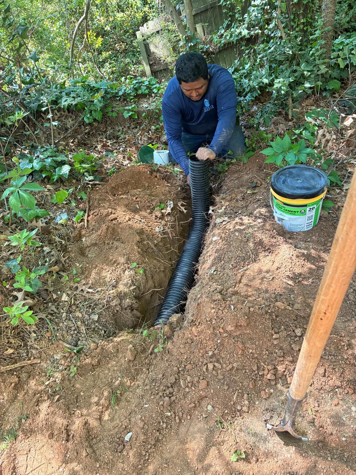 Person installing a black pipe in a dirt trench outdoors, with tools and a bucket nearby
