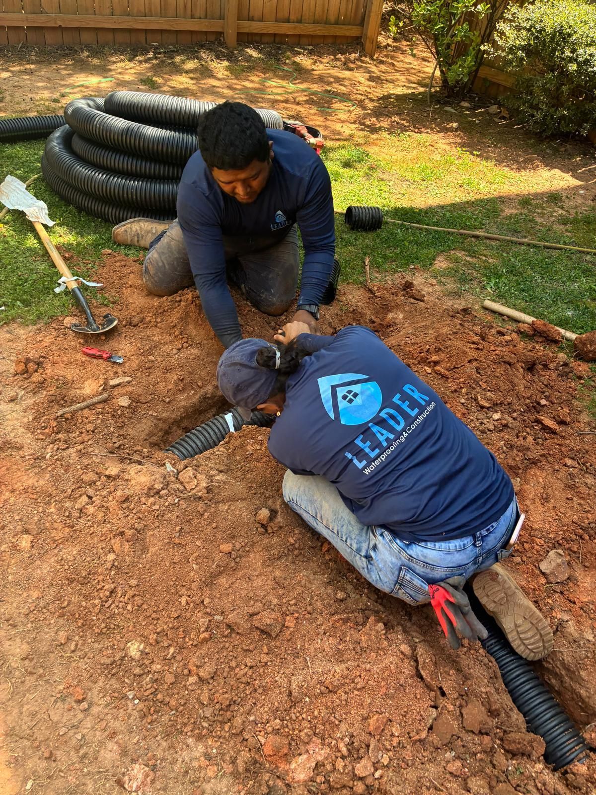 Two workers kneel on dirt, connecting black corrugated pipe in a yard near stacked hose rings.