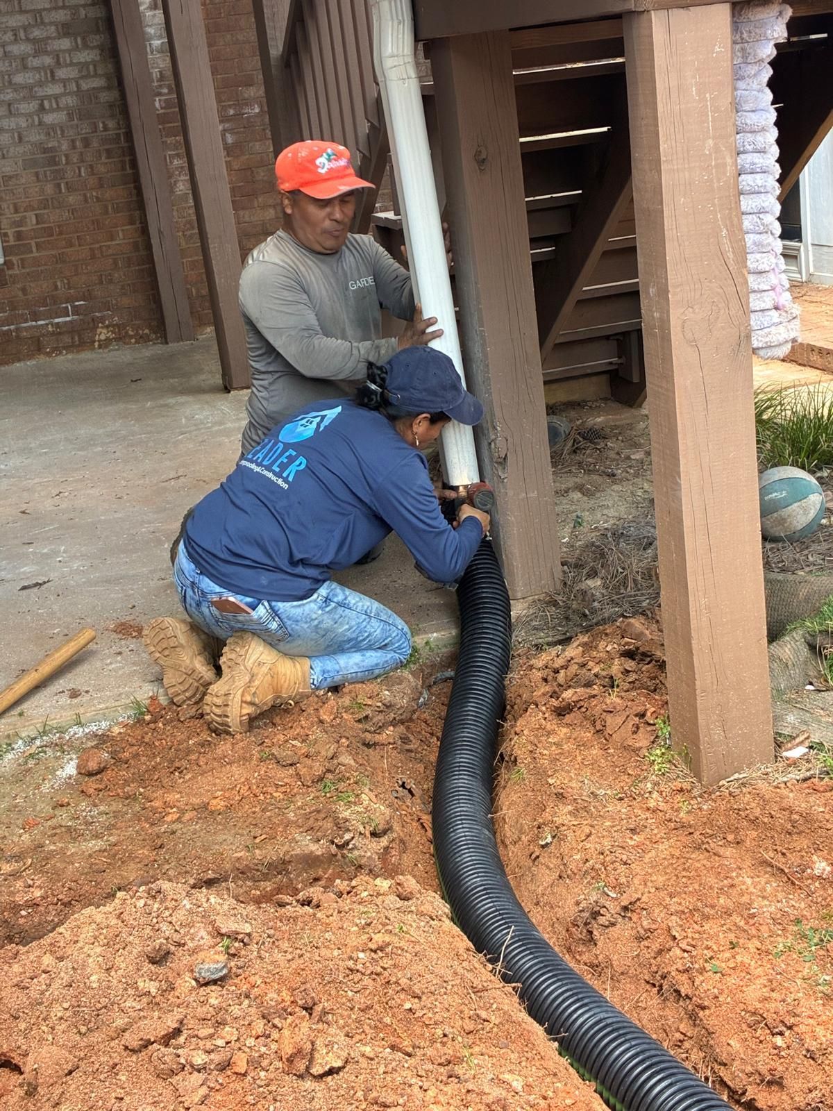 Two workers installing a black drainage pipe beside a wooden post on a dirt construction site.