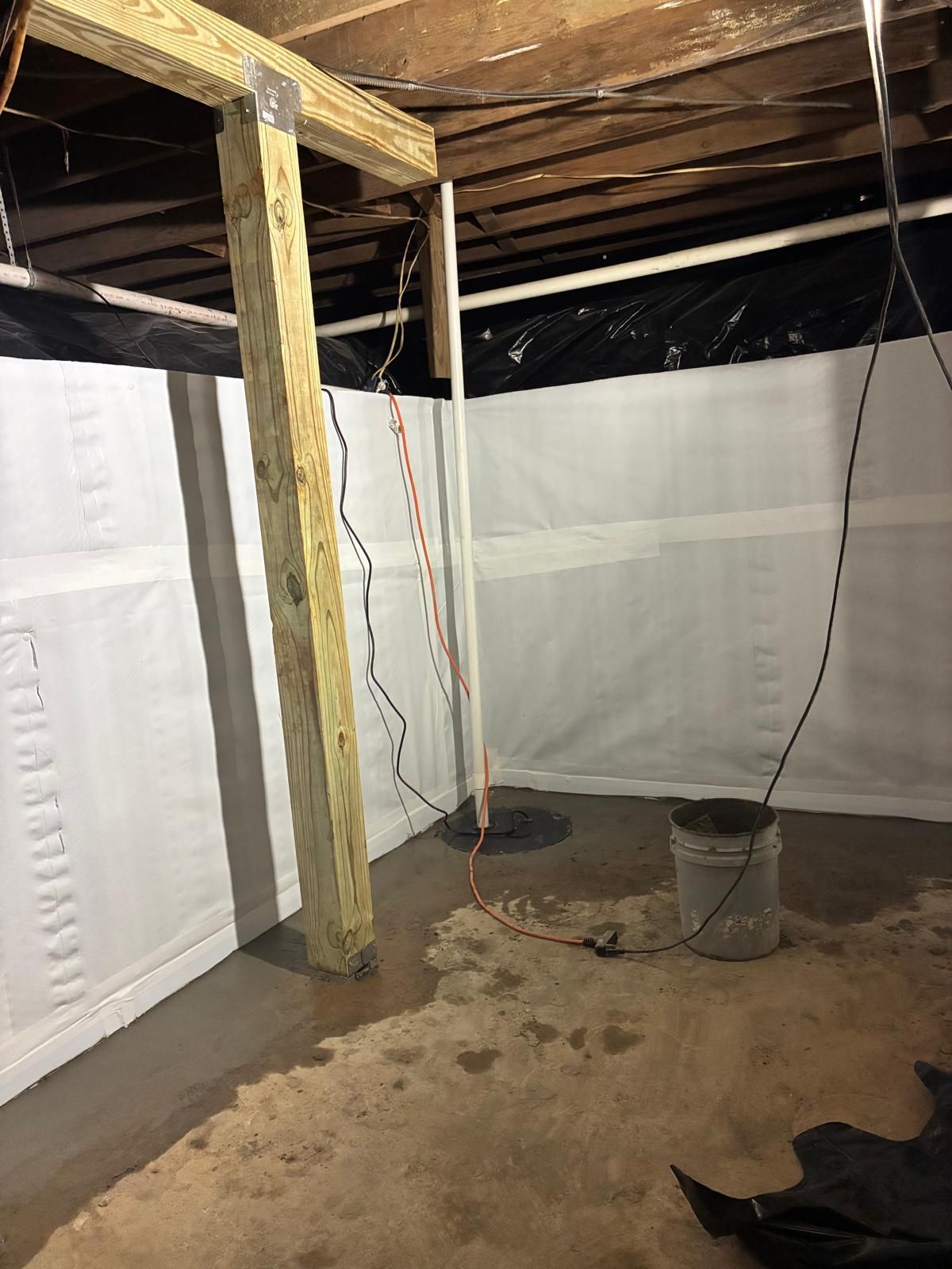 Partially finished basement with exposed wooden beams, plastic wall vapor barriers, and a bucket on the floor