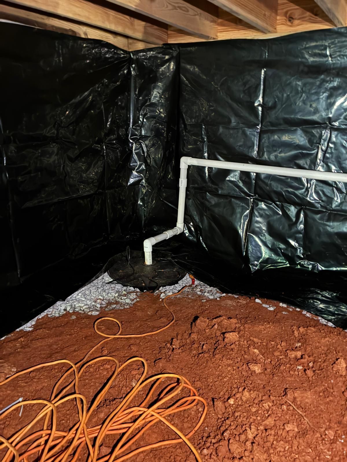 Basement crawlspace with black plastic walls, white PVC pipe, red dirt floor, and orange extension cord