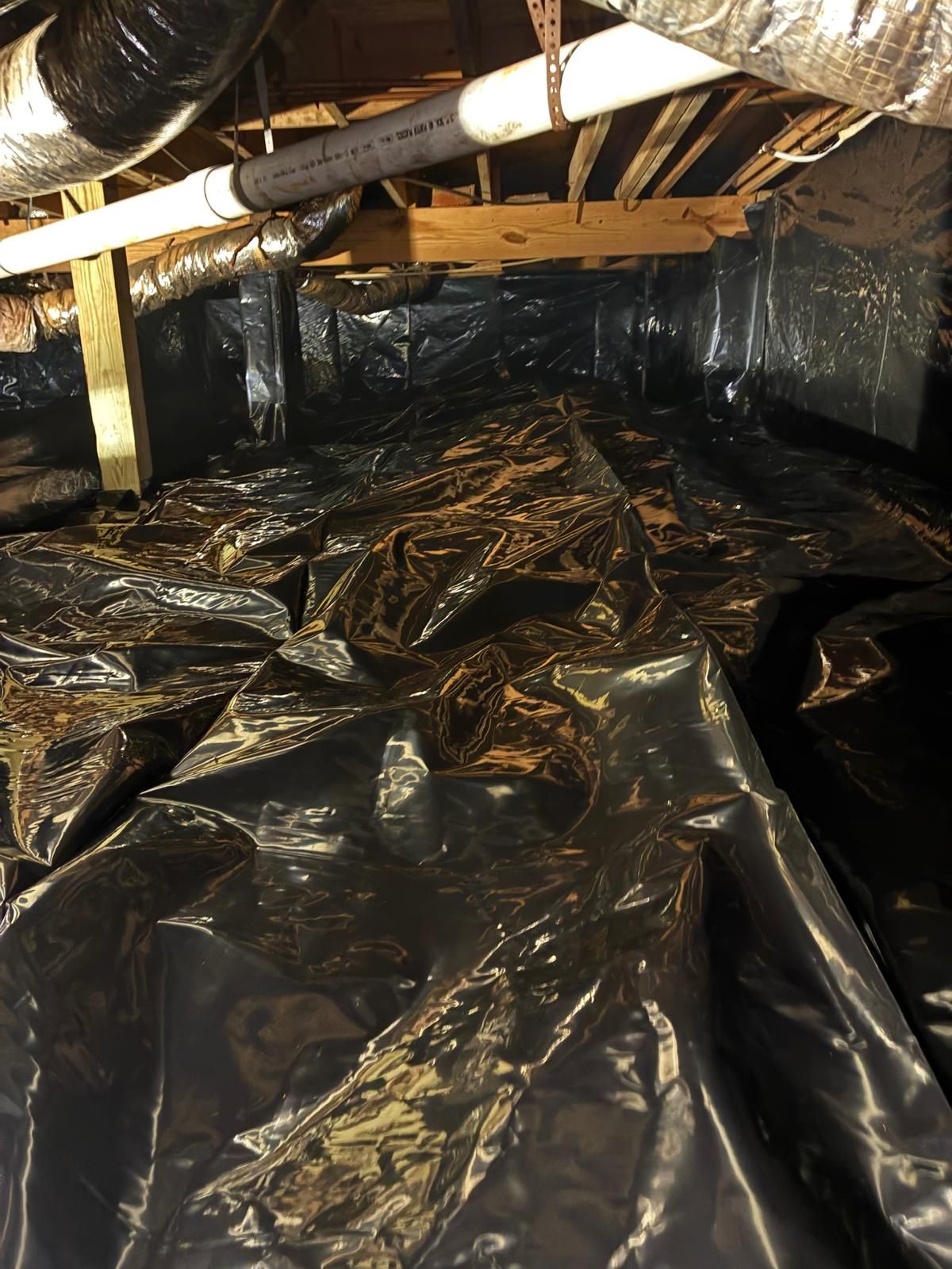 Insulated crawlspace with black plastic vapor barrier and exposed pipes and ductwork