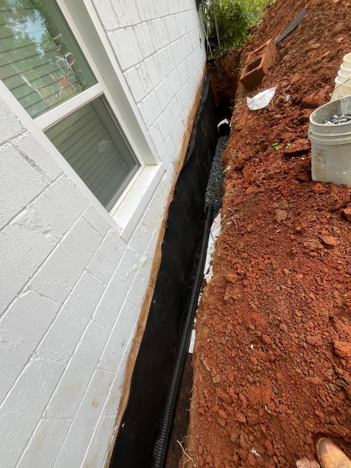 Narrow trench beside a white house foundation, lined with black drainage material and red dirt.