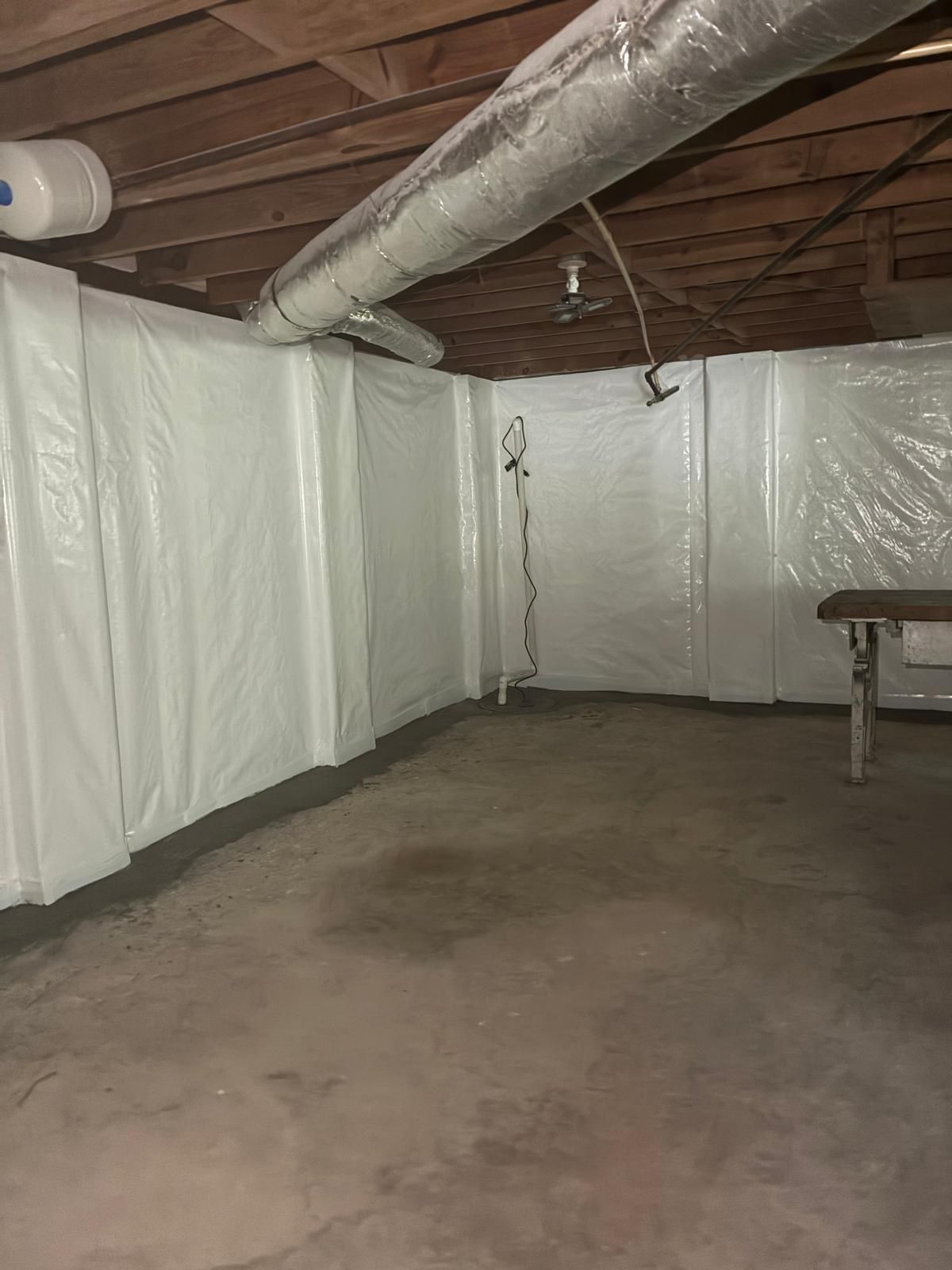 Unfinished basement with exposed ceiling beams, plastic wall sheeting, concrete floor, and a workbench.