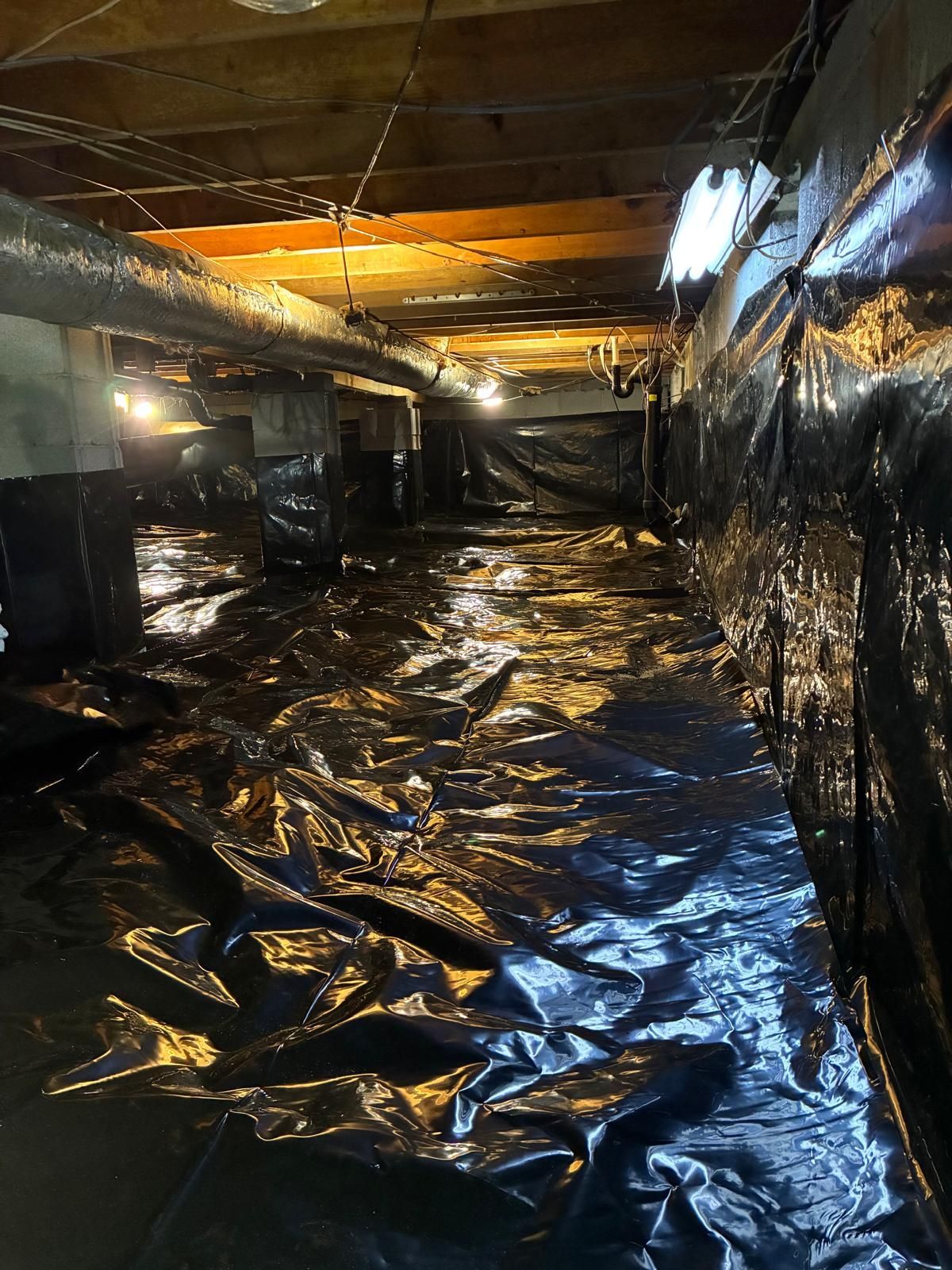 Dark basement crawlspace lined with black plastic sheeting and overhead pipes, lit by a warm work light.