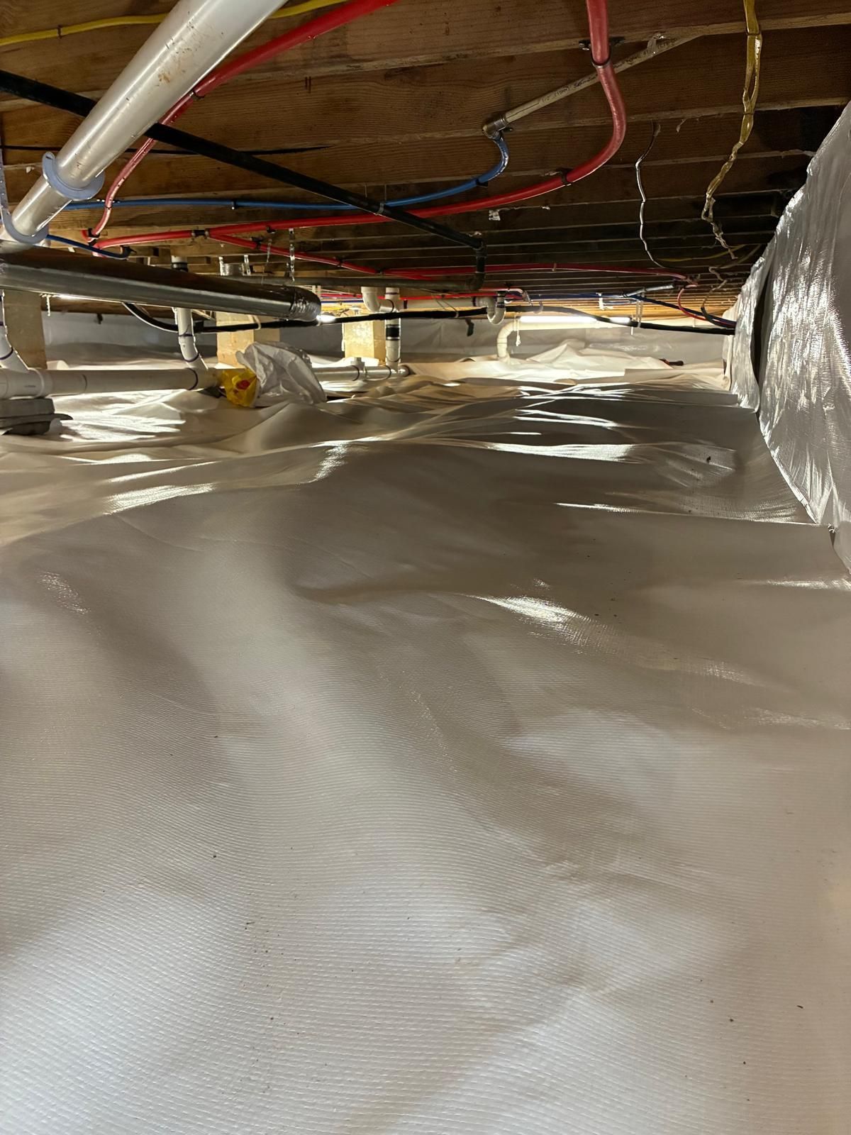 Crawlspace under a house with insulated pipes and a white vapor barrier on the ground.