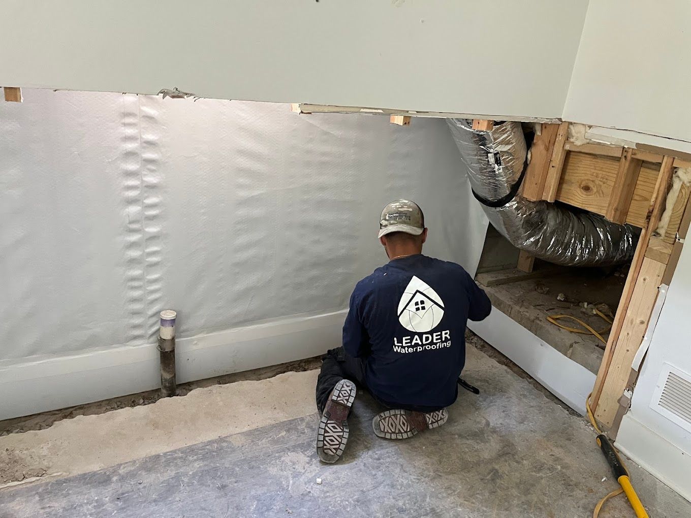 Worker kneeling in a basement, applying white wall barrier near exposed framing and ductwork.