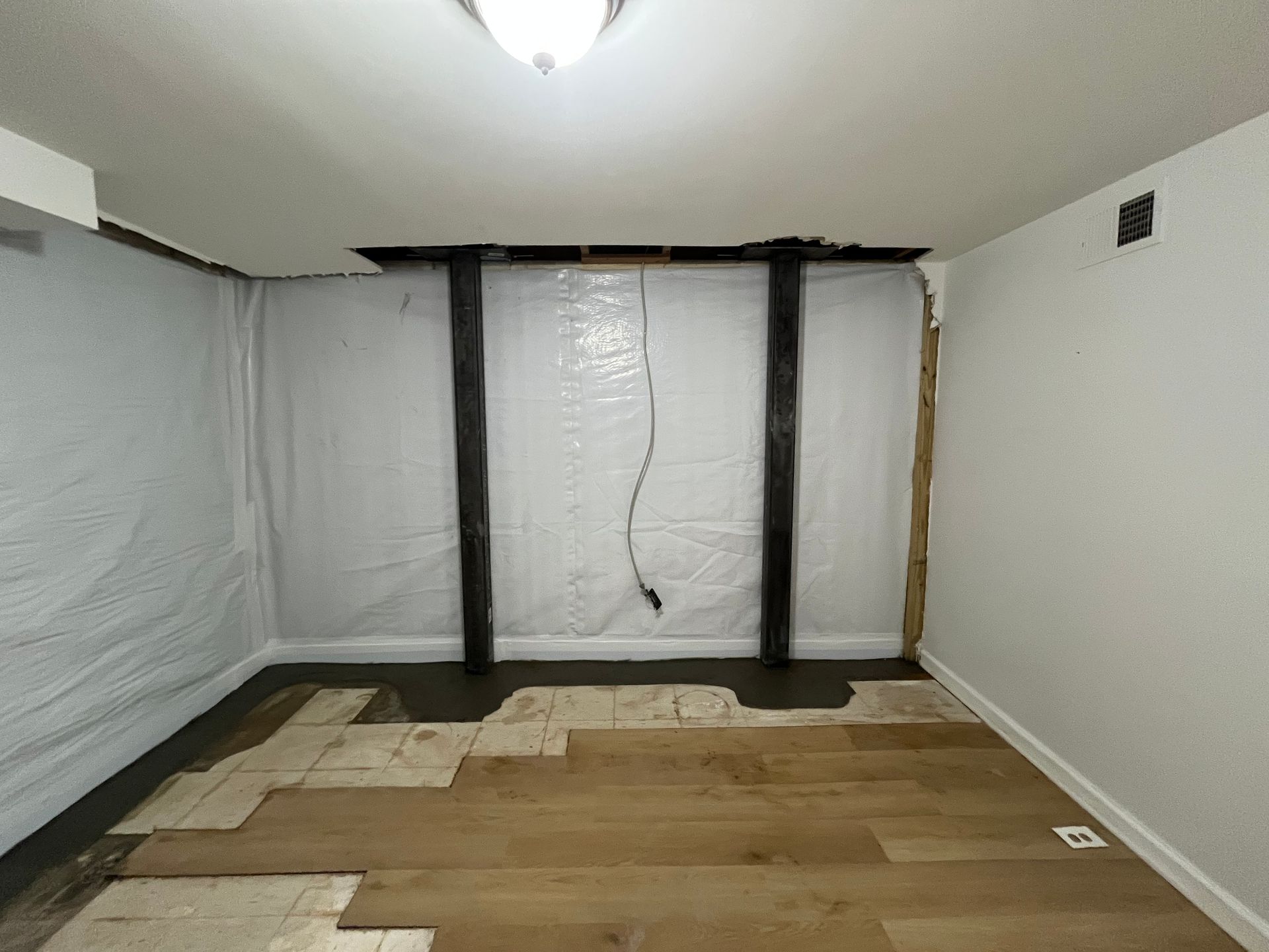 Empty room with unfinished hardwood floor, white walls, and plastic sheeting covering the back wall.