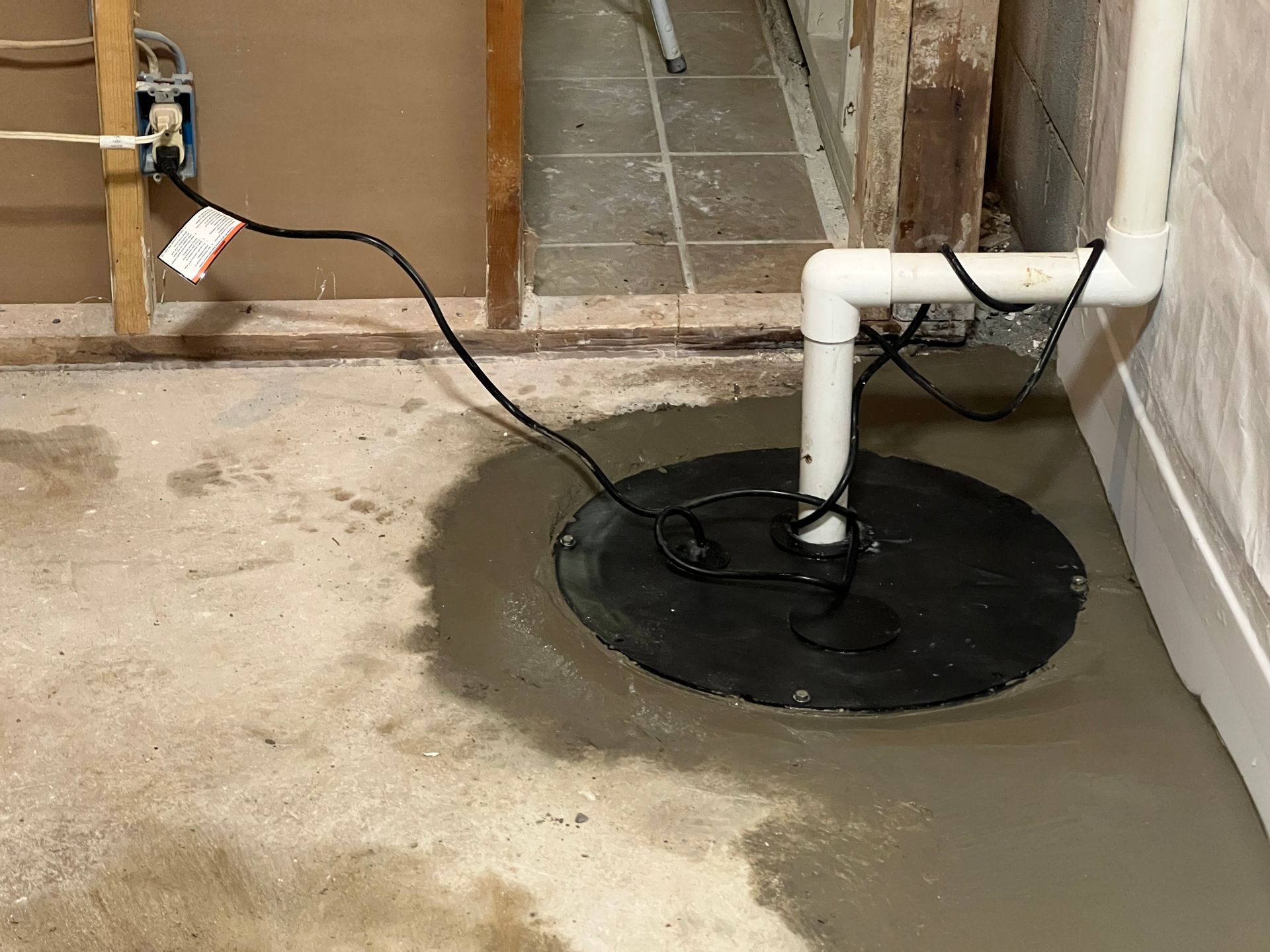 Water meter in a damp basement, with white PVC pipes and a black sump cover on the floor.