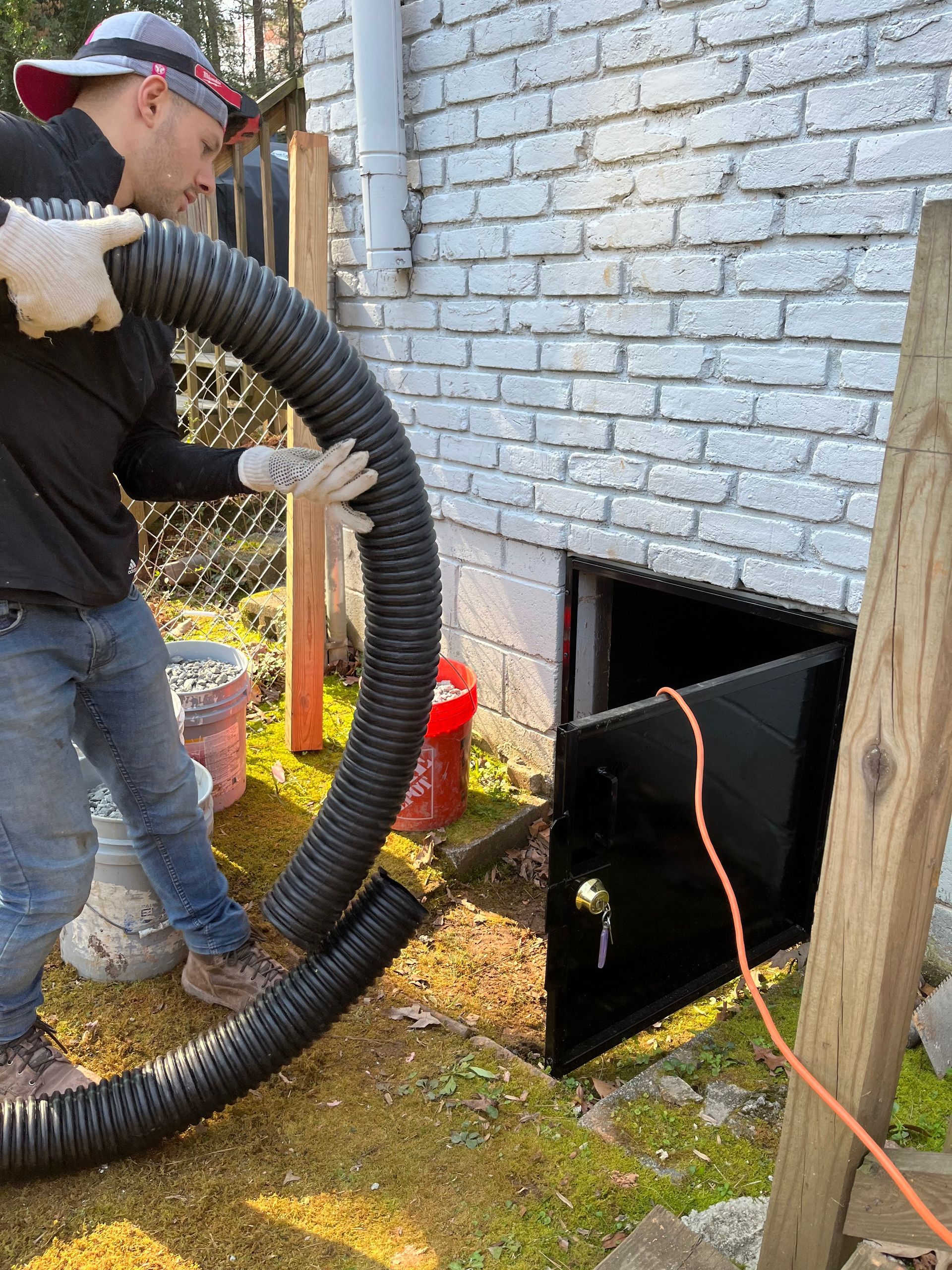 Worker vacuuming debris from a basement vent with a large hose beside a brick foundation wall