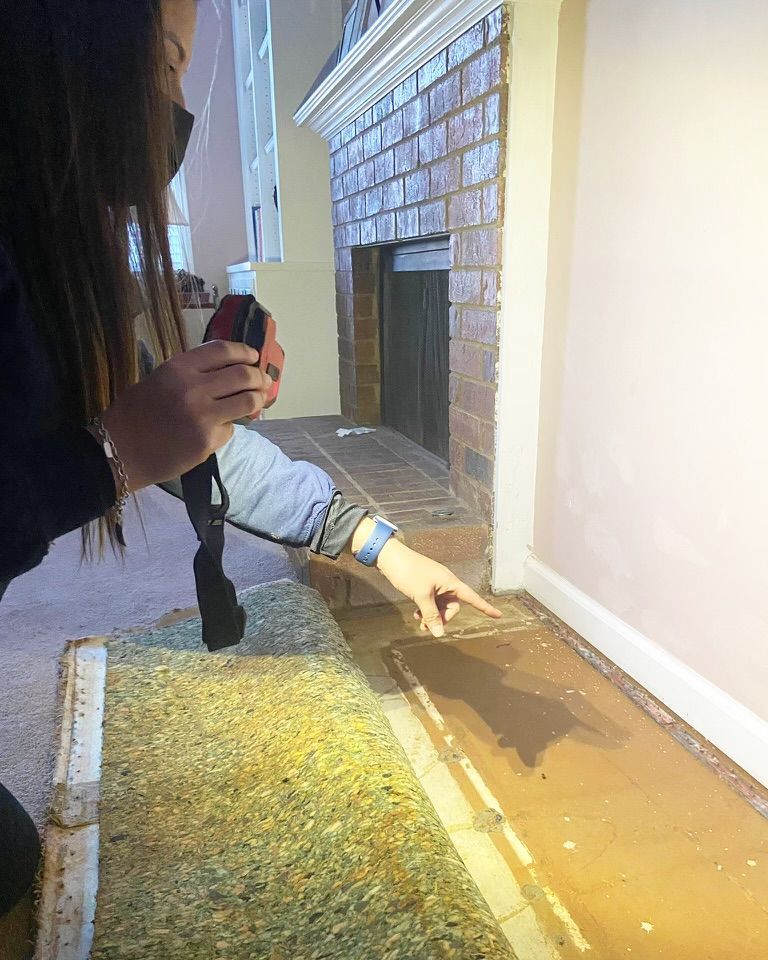 Person points at muddy water pooled along a hallway wall near a brick fireplace.