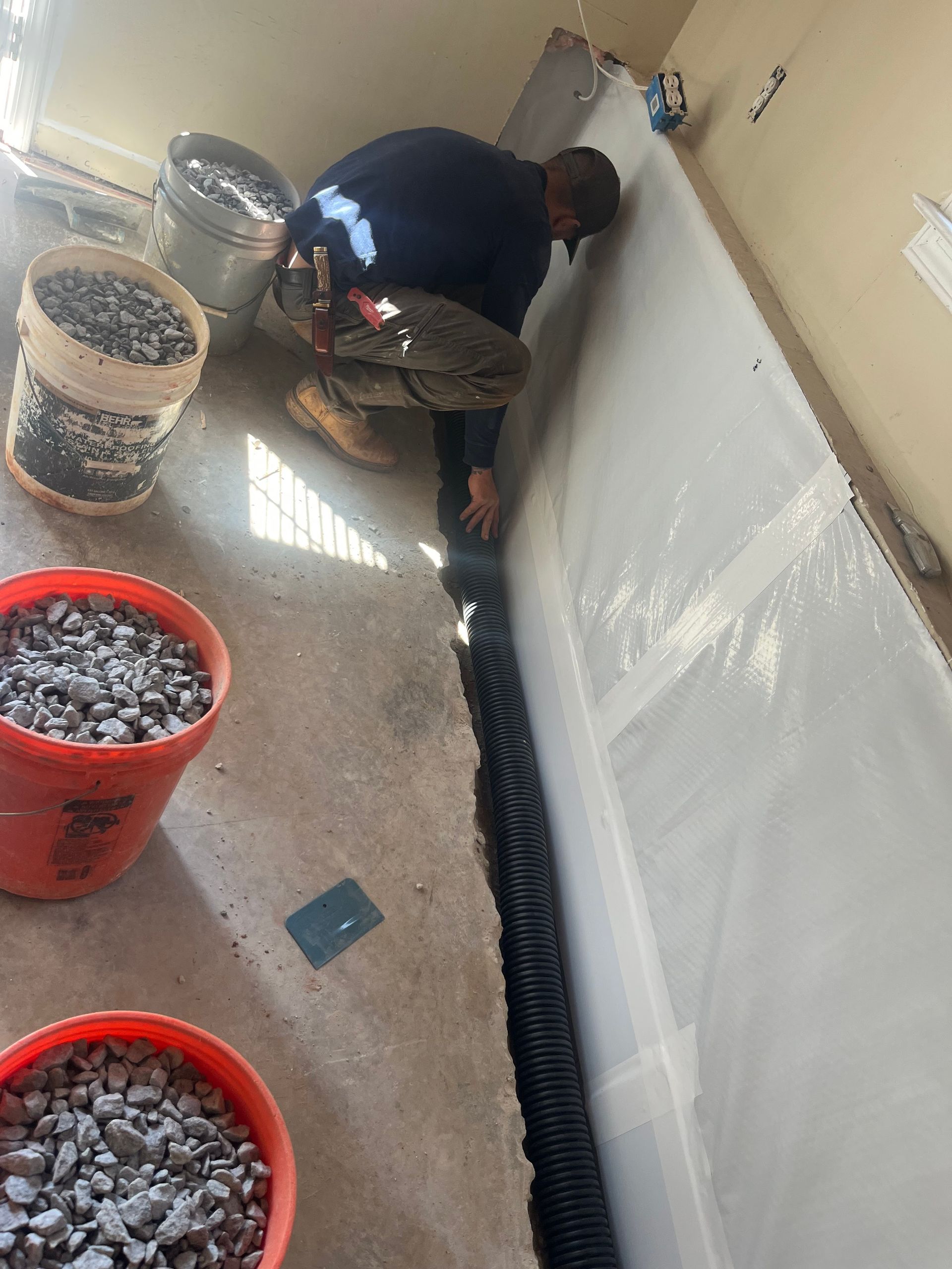 Worker installing drainage pipe along wall beside buckets of gravel in a narrow room