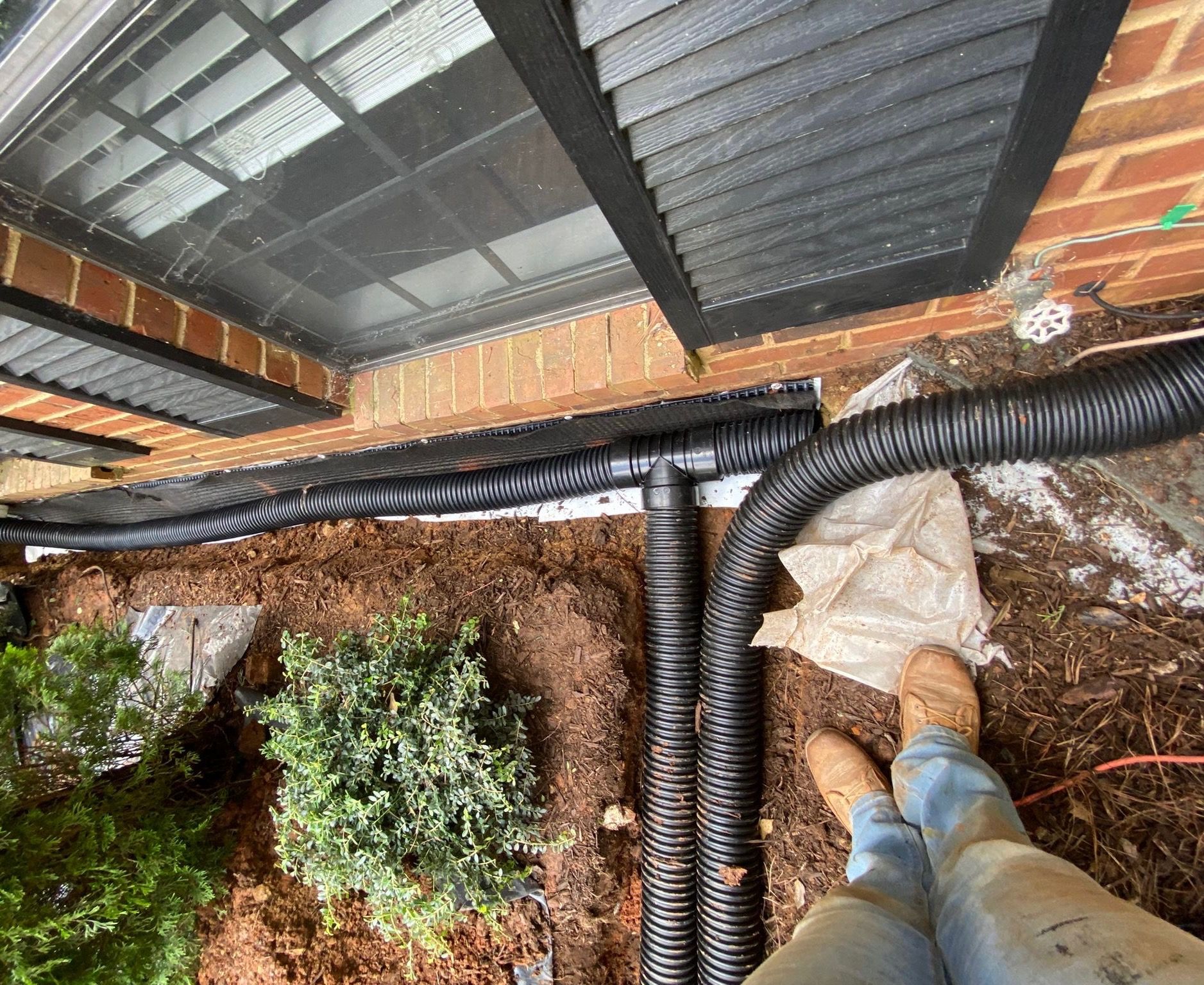 Worker standing beside black corrugated pipes on a rooftop or terrace garden, looking down at soil beds.