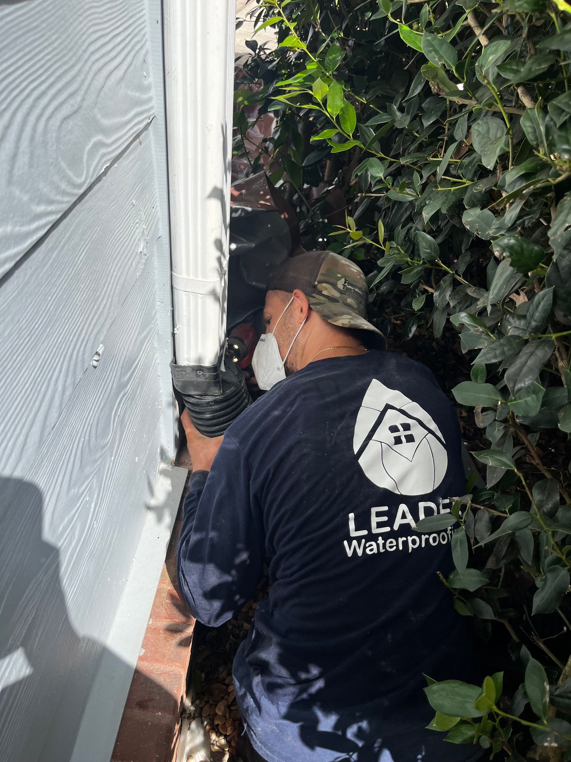 Worker in a black jacket inspects a house exterior beside dense bushes.
