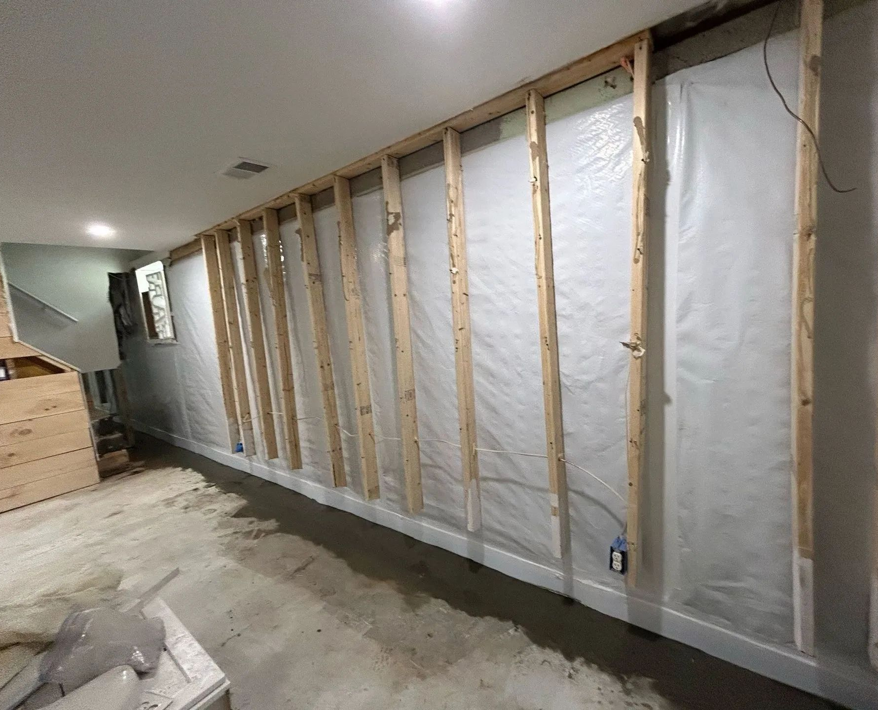 Unfinished basement wall framed with studs and insulated with white vapor barrier.
