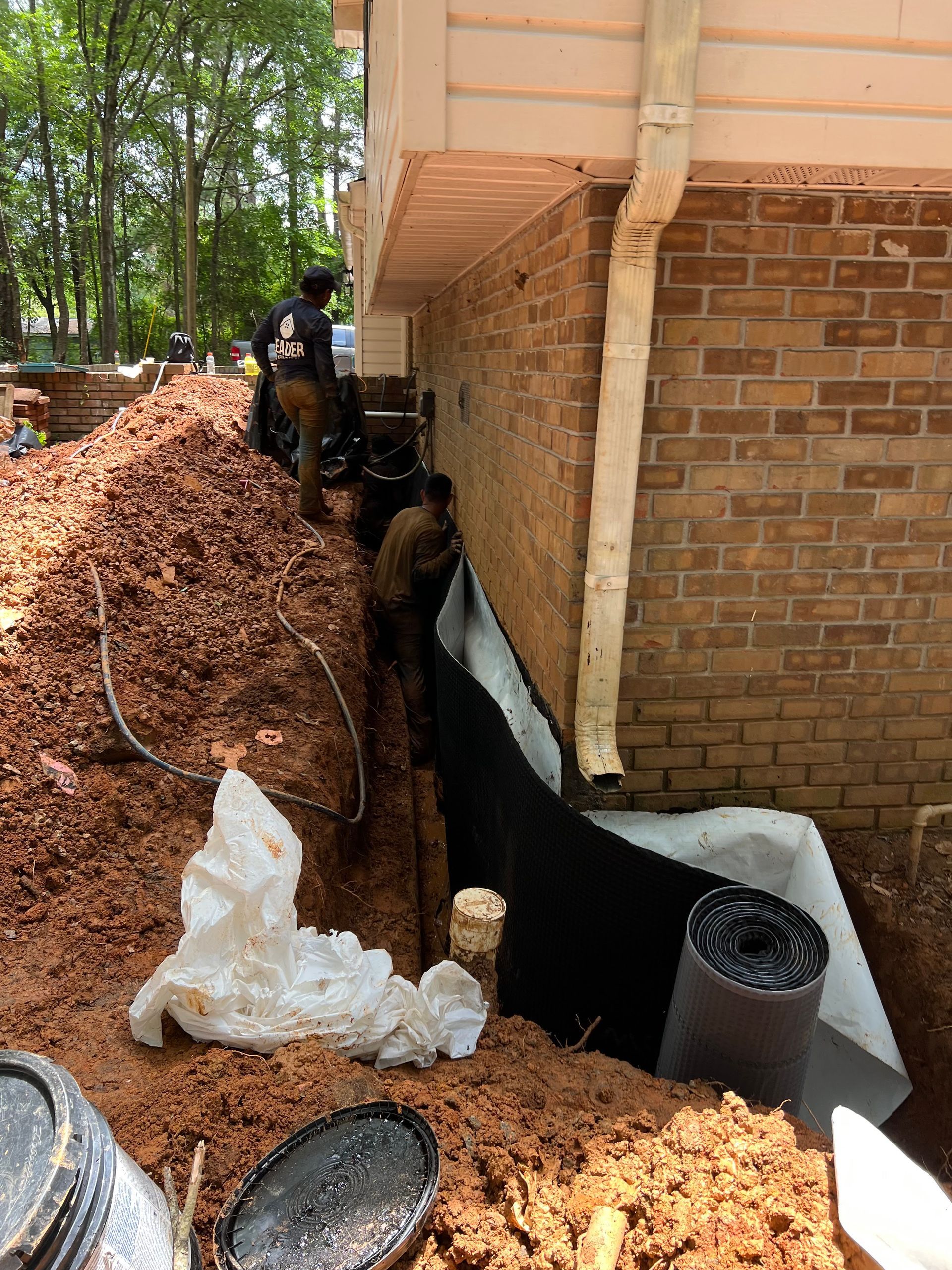 Foundation repair trench beside brick house, with black waterproofing, dirt piles, and construction equipment