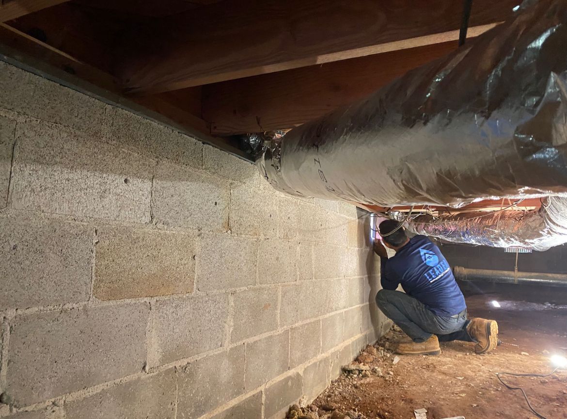 Worker crouching in a crawlspace beside a cinderblock wall and exposed ducting, inspecting insulation.