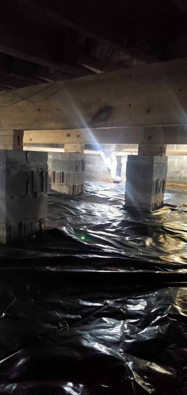 Damp crawl space under a house with plastic sheeting on the ground and water reflecting light between beams