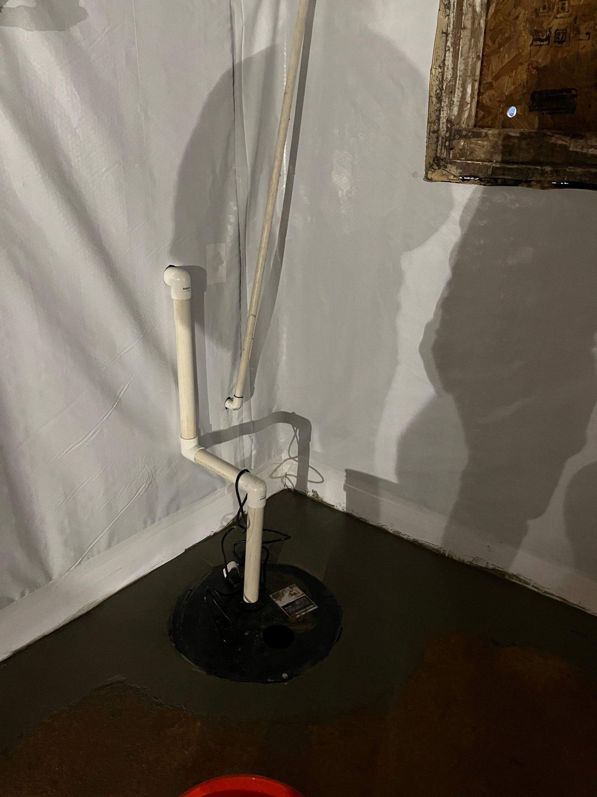 Black sink drain in a white wall corner with exposed pipes and dark floor liquid