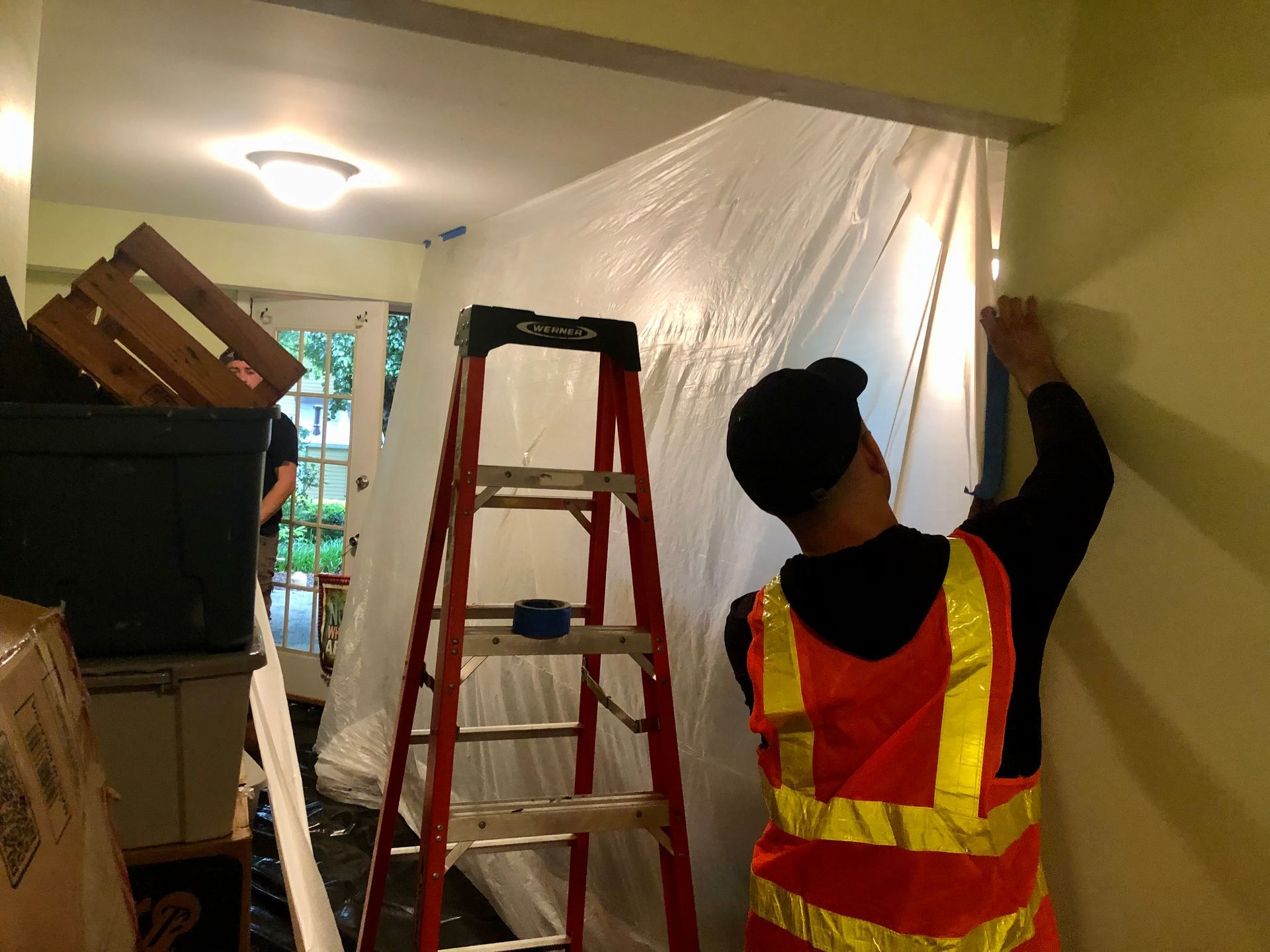 Worker in orange safety vest painting a room, with ladder and plastic sheeting protecting the area.