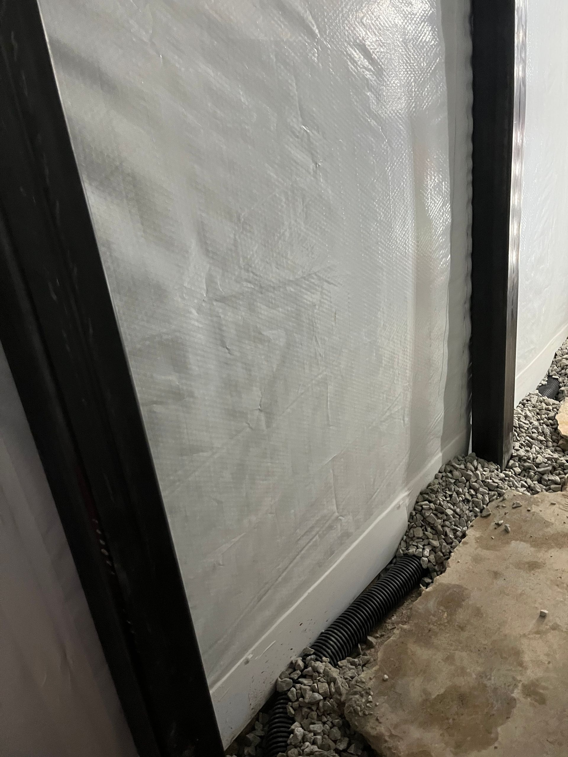 White tarp or wall panel beside a black frame, with gravel and a wet concrete path below.
