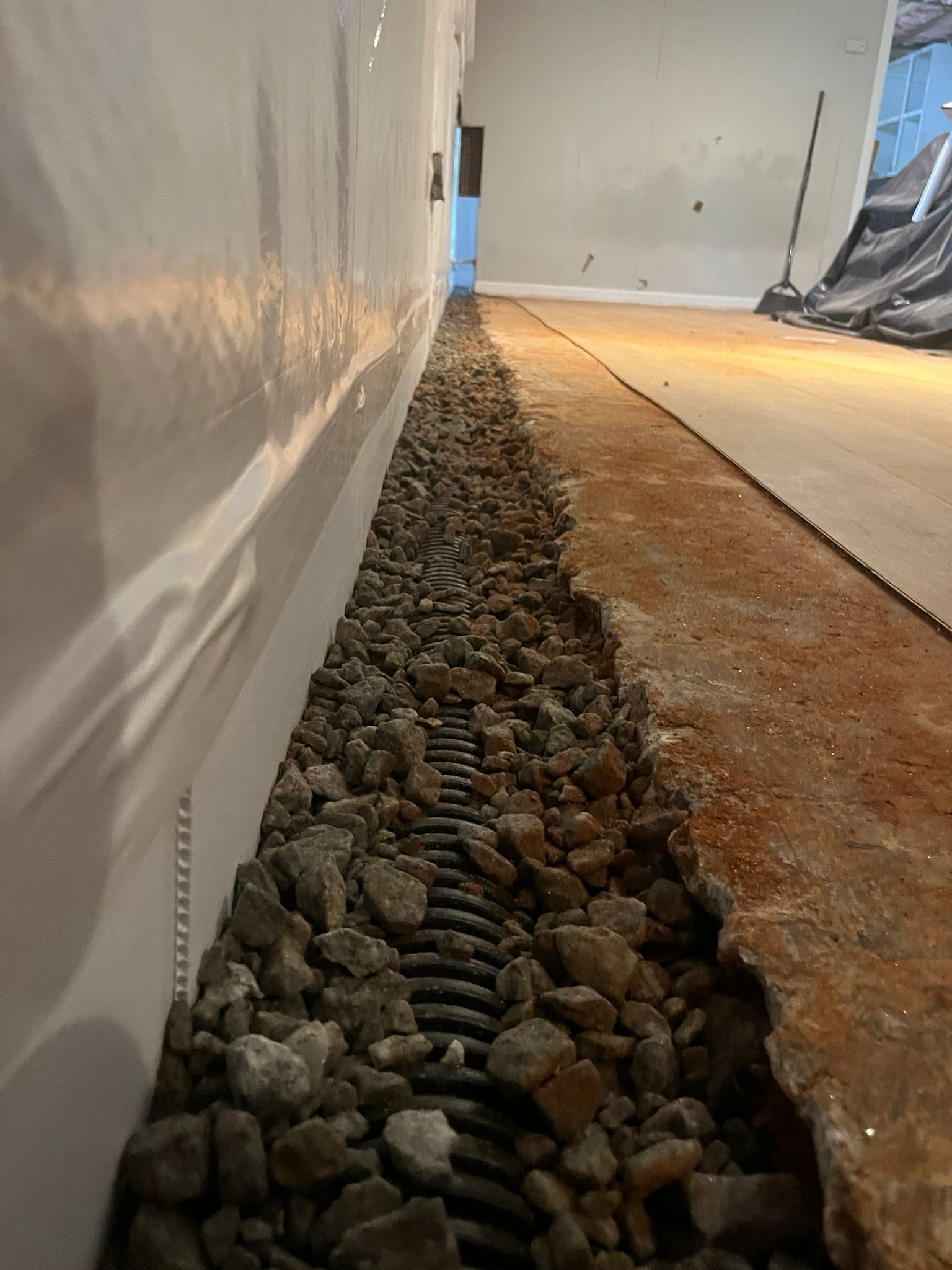 Narrow gravel drainage strip beside a white wall and concrete floor in an unfinished interior.