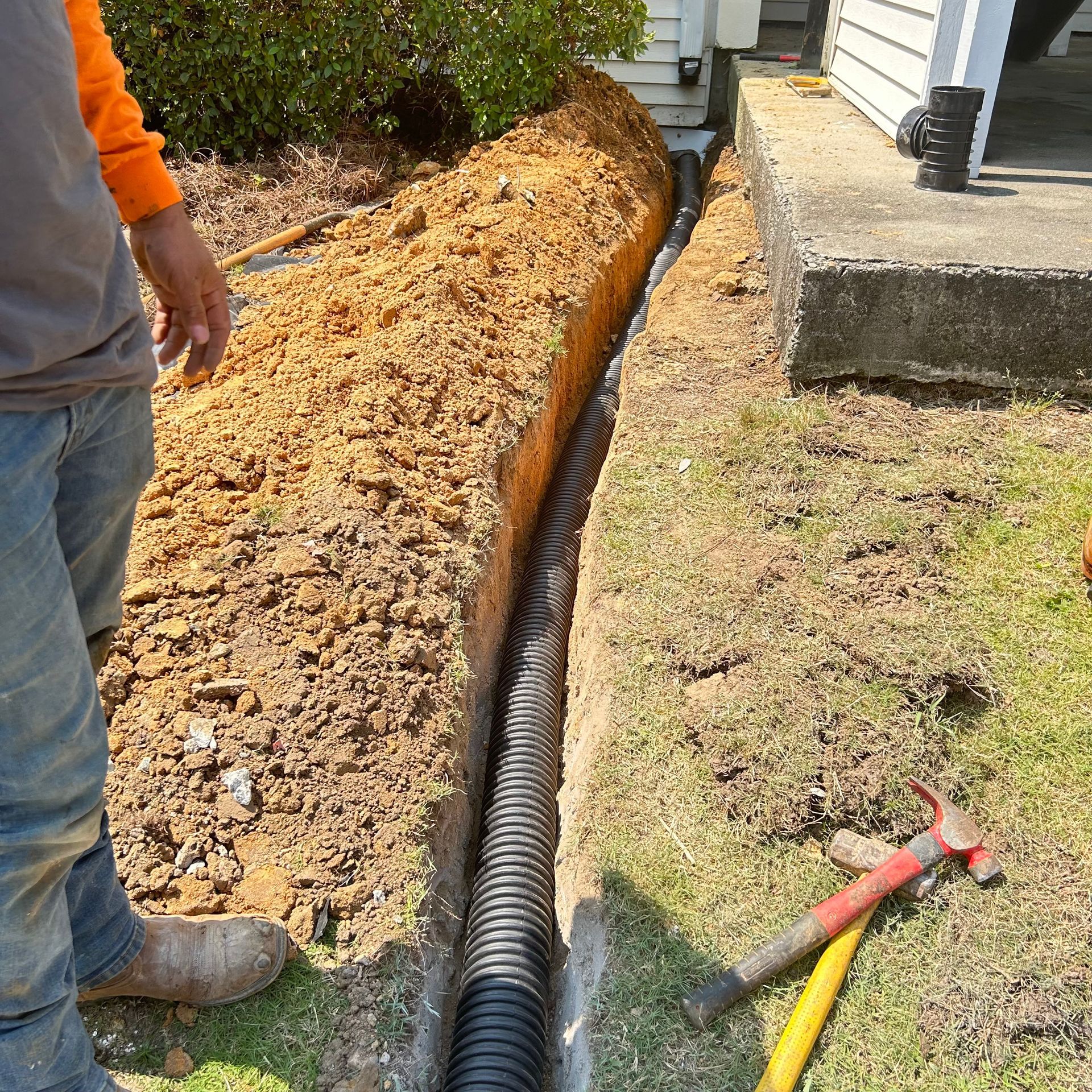 Trench beside a house with black drainage pipe and tools in the dirt; worker stands nearby.