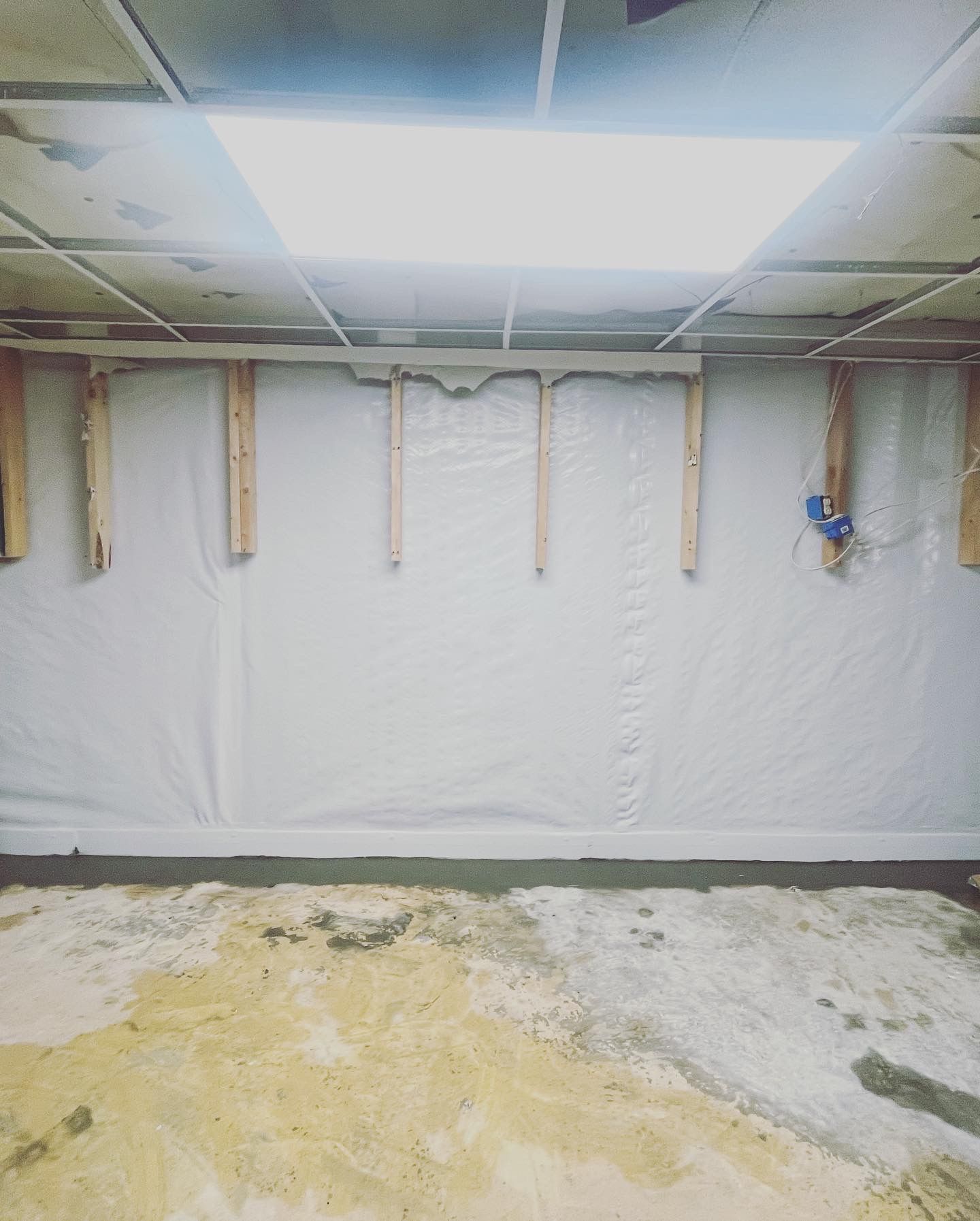 Basement wall under renovation with exposed studs, plastic sheeting, and debris-covered concrete floor