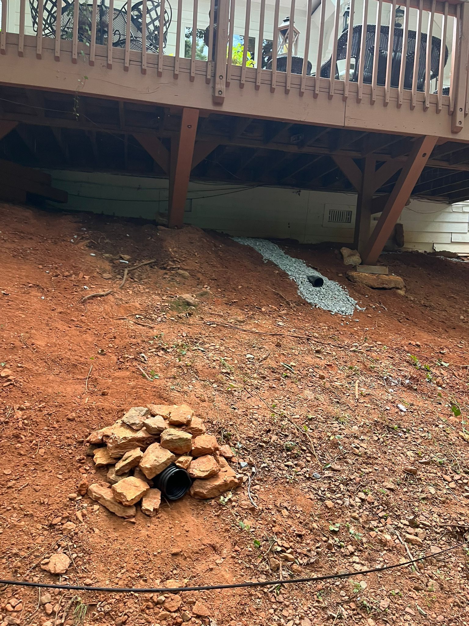 Backyard under a deck with red gravel, a small stone fire pit, and a white drainage strip on a slope