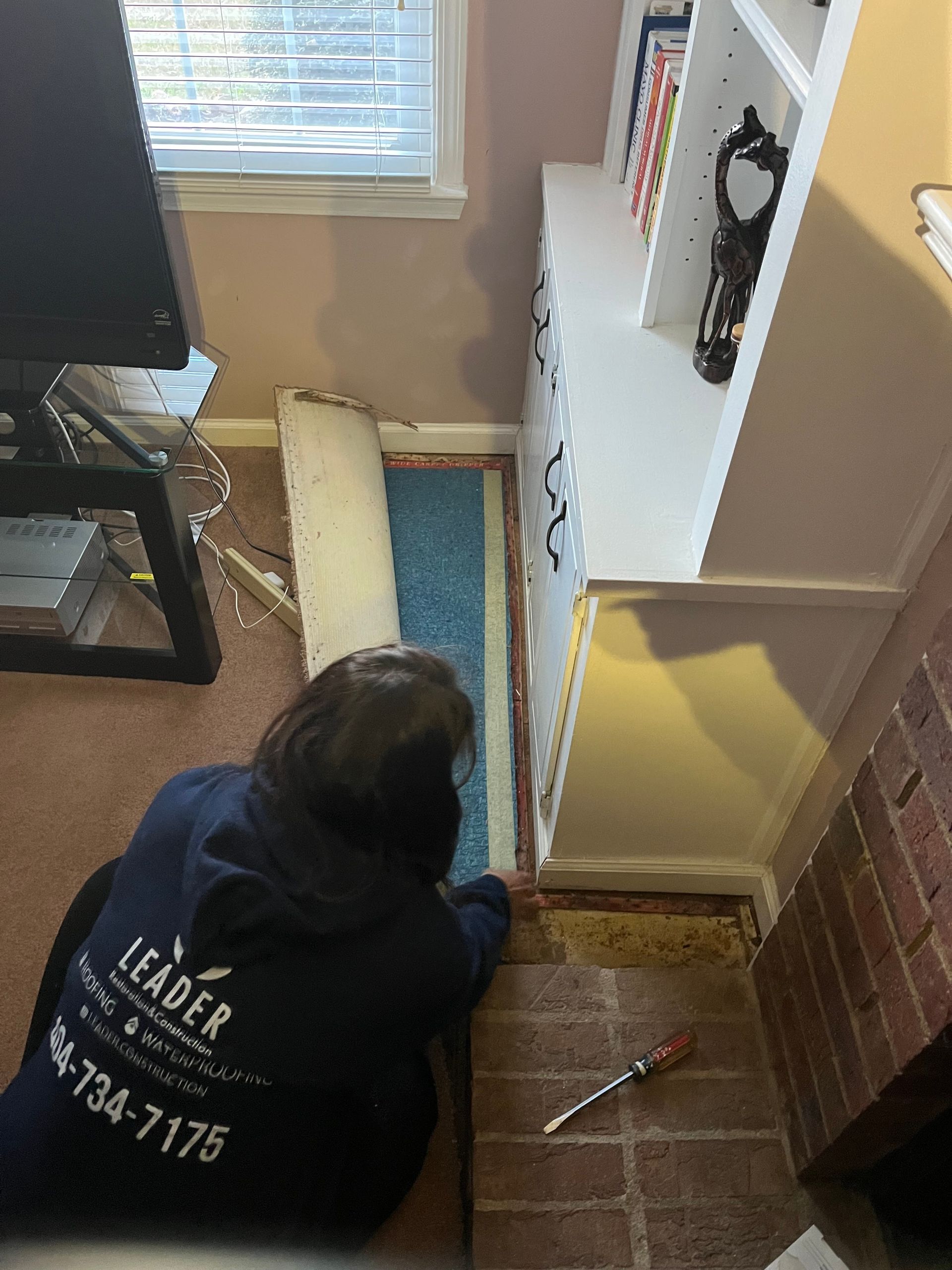 Person kneeling by a window, installing flooring beside a white cabinet in a small room.