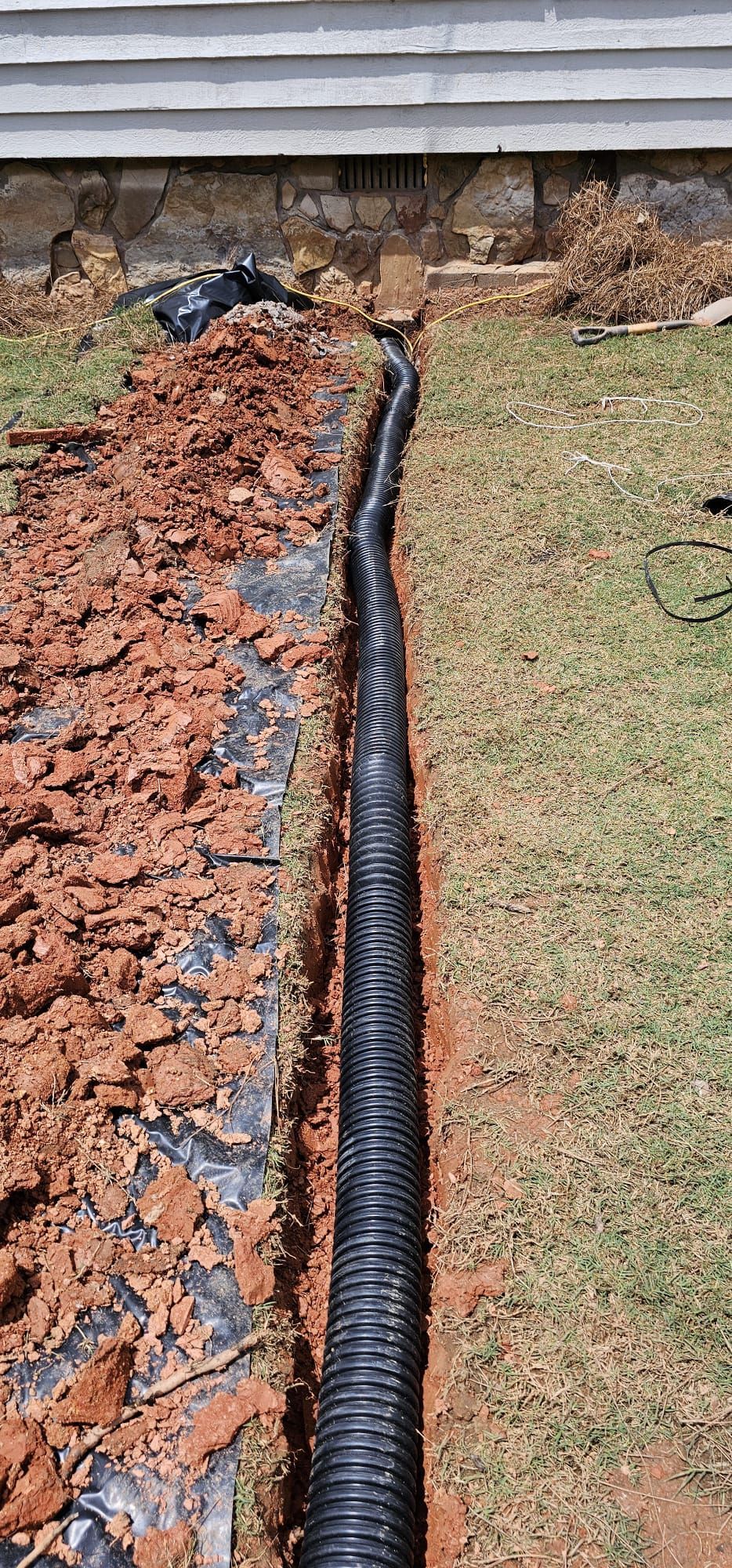 Black corrugated drain pipe laid in a narrow trench beside a house foundation with fresh soil and grass edge