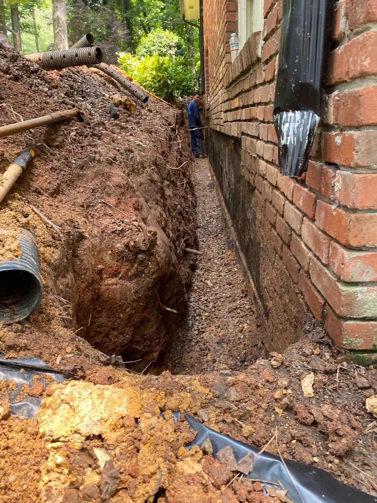 Narrow trench beside a brick house, with muddy soil piles and a worker in the background